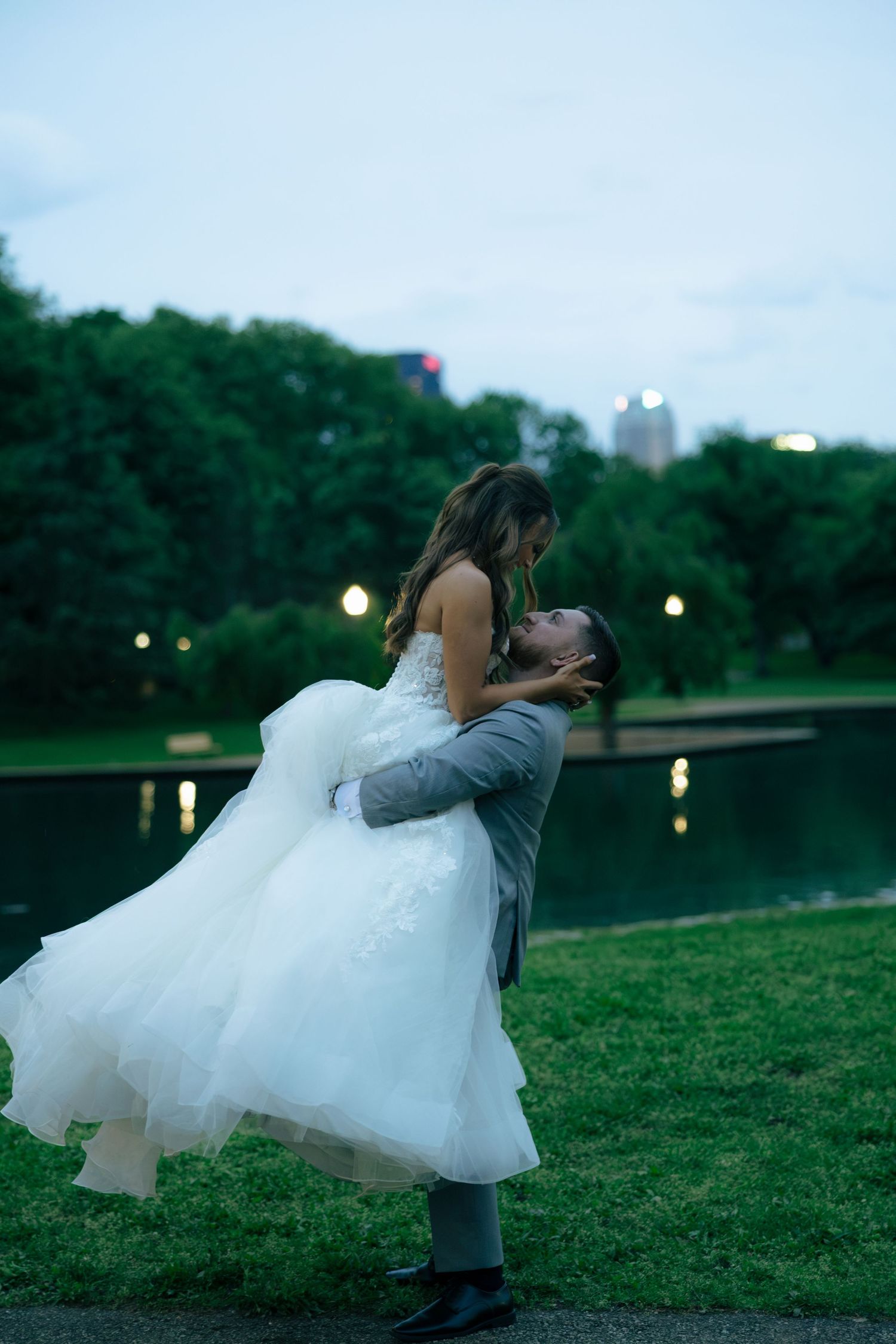 Groom lifts bride in white ballgown by a park lake at dusk during romantic wedding photo session.