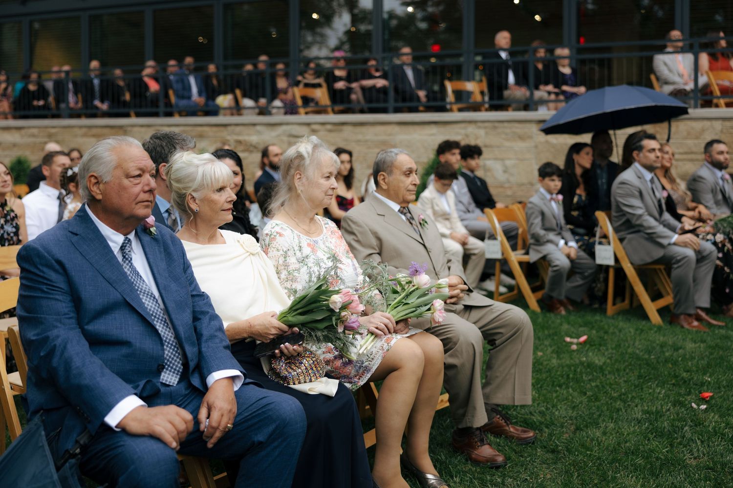 Wedding guests seated in wooden chairs at outdoor ceremony, woman holding white flowers in front row.