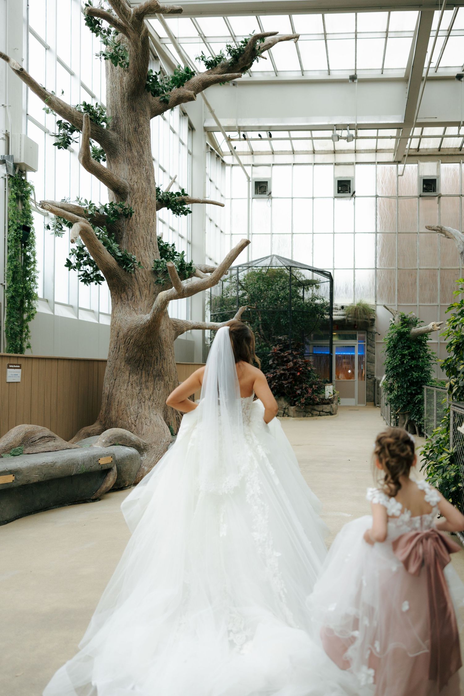 Bride in white gown with long veil walks with flower girl in pink dress through elegant indoor garden venue.