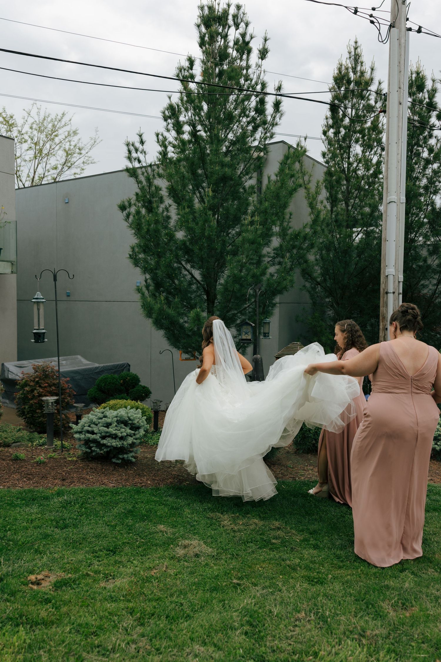 Bridesmaids in mauve gowns help carry bride's white wedding dress train across garden lawn outdoors.