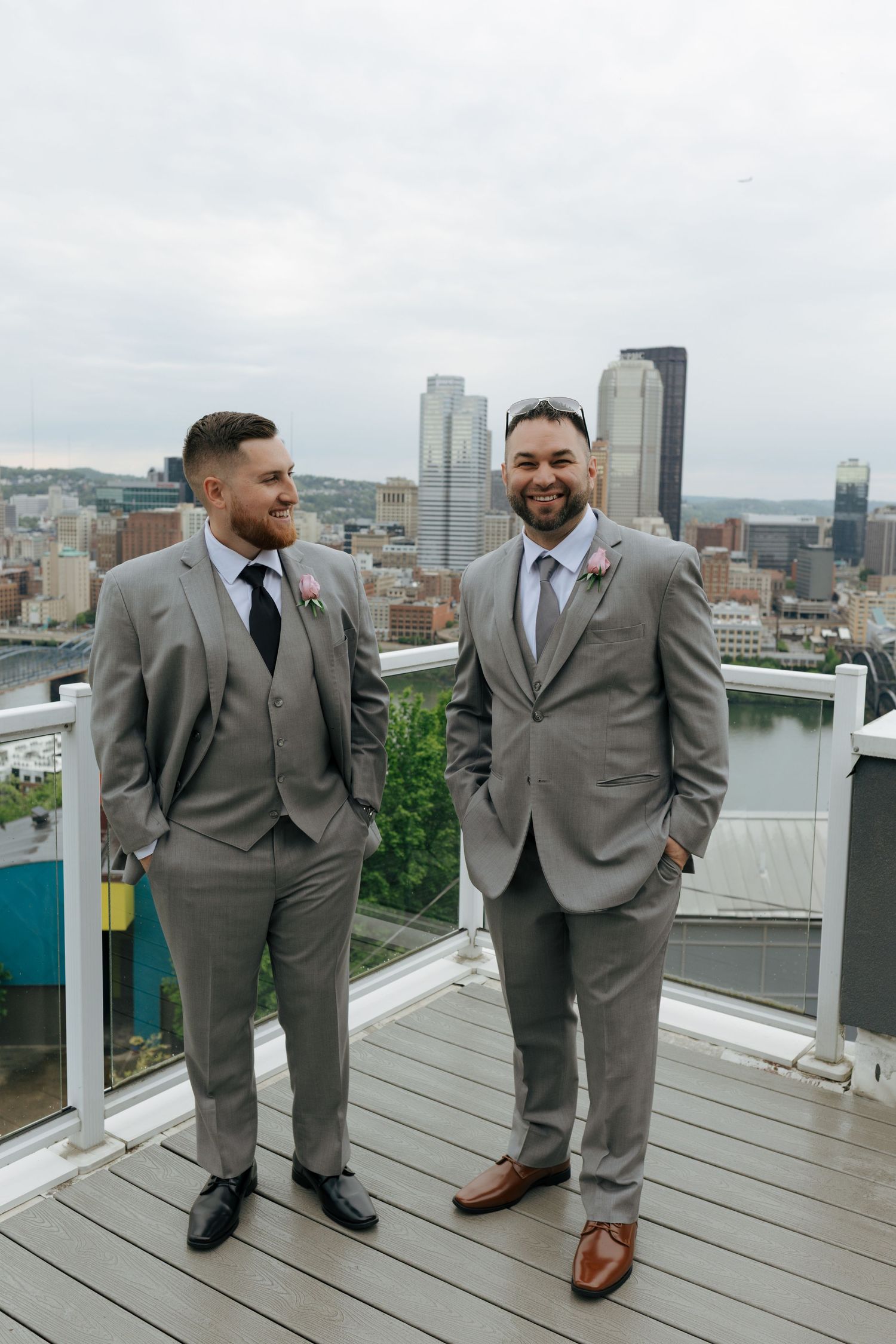 Two groomsmen in matching gray suits smile together on a rooftop overlooking the Pittsburgh city skyline.