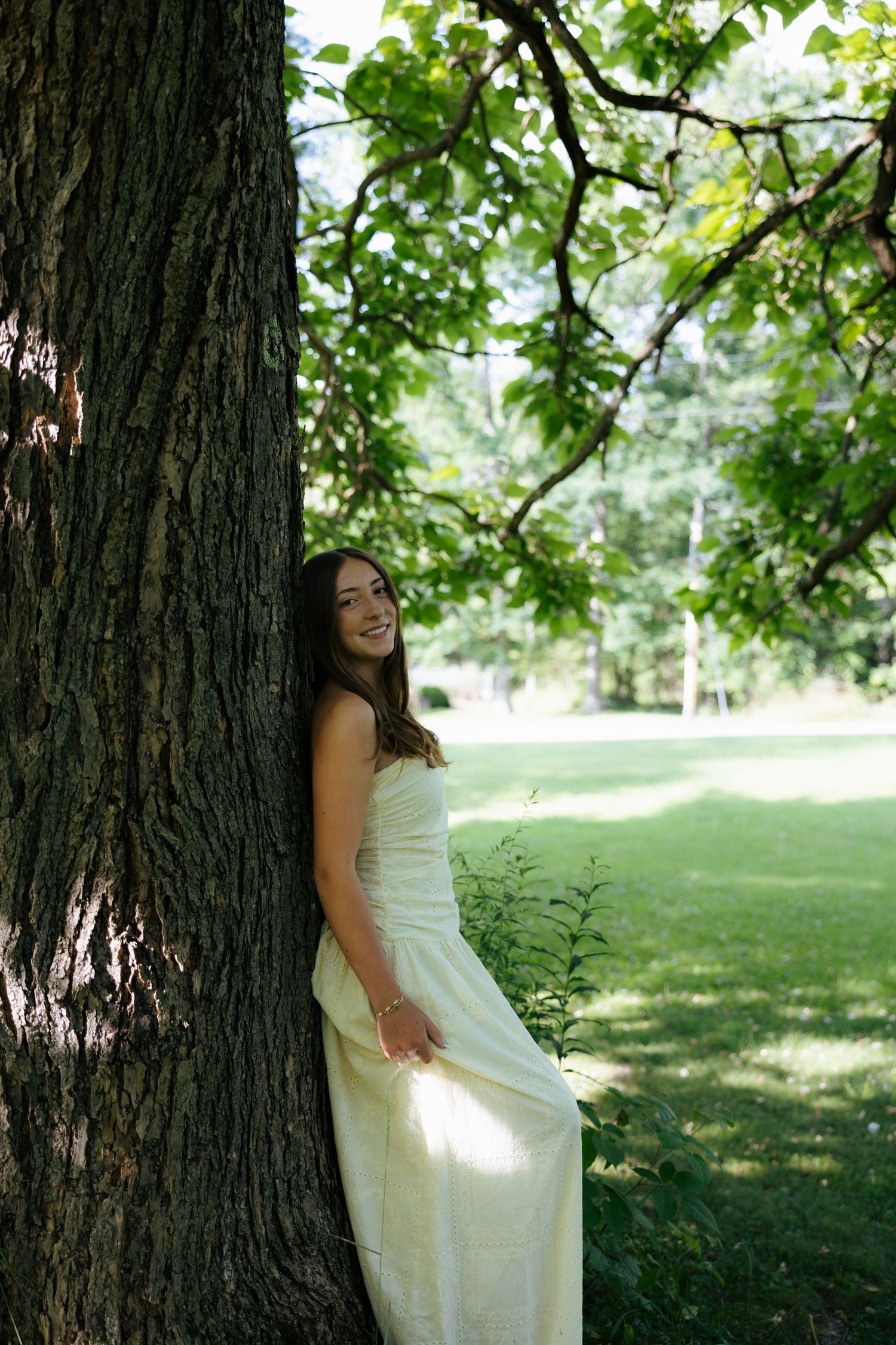 Young woman in white dress leaning against large tree trunk in lush green park, smiling.
