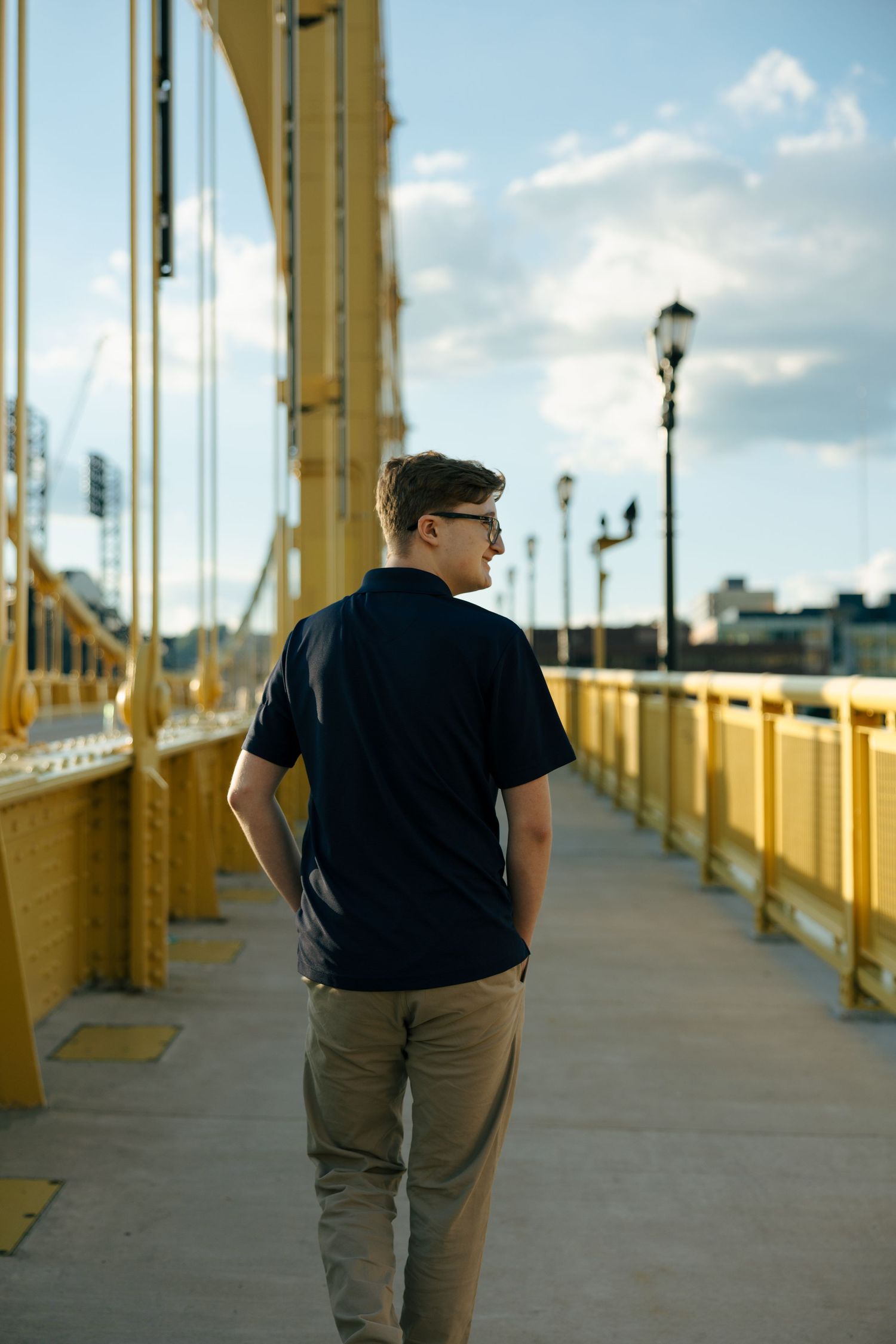 Young man in black t-shirt and khaki pants walking on a yellow bridge pedestrian path on a sunny day.