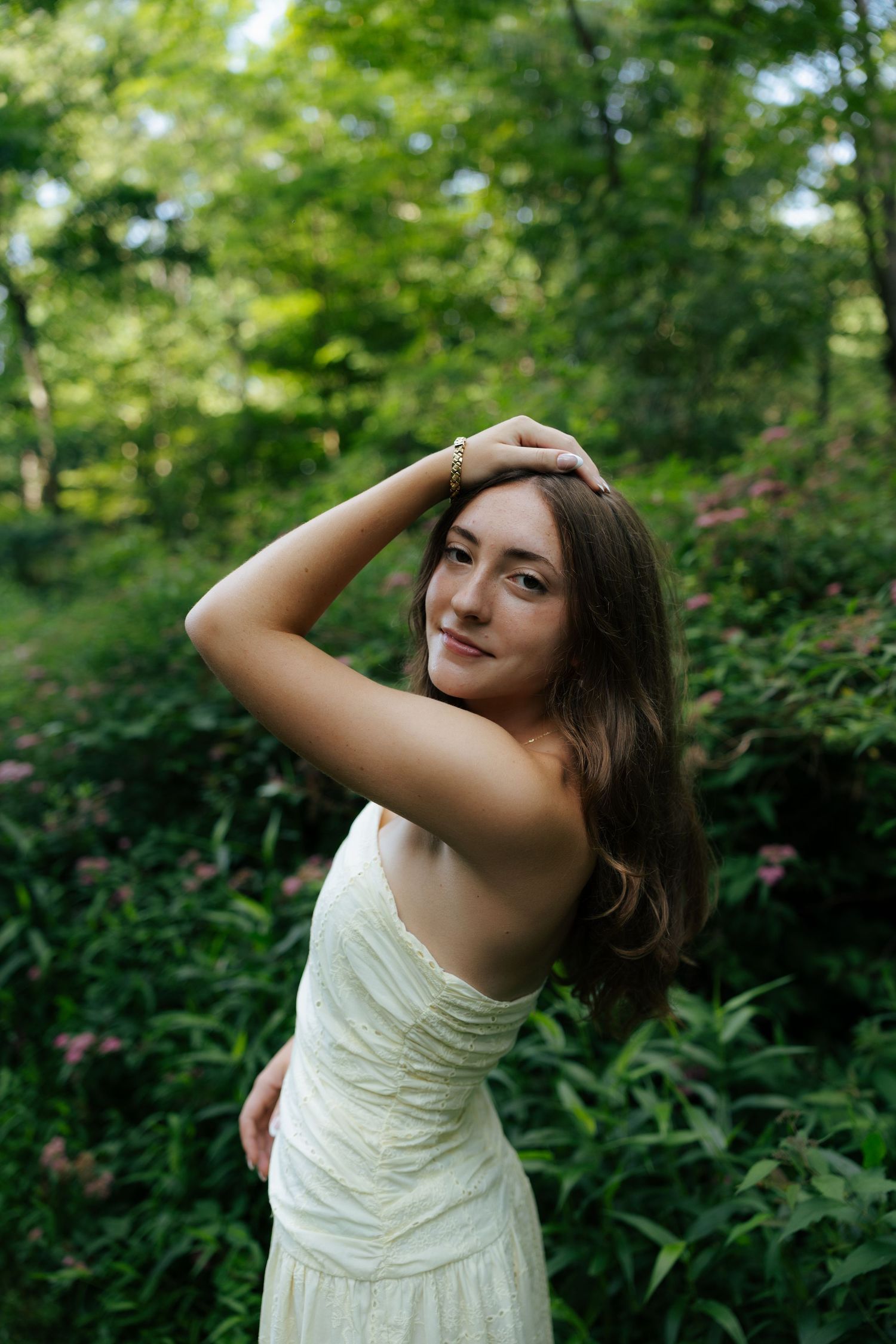 Young woman in white dress posing gracefully among lush green foliage and pink flowers.