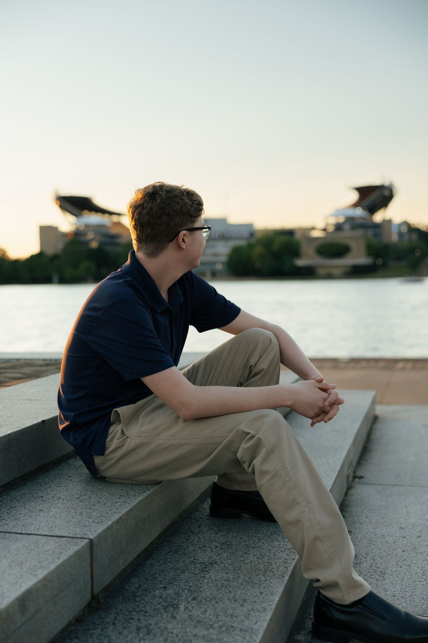Teen boy in navy polo sitting on stone steps overlooking a river with a stadium in the background.