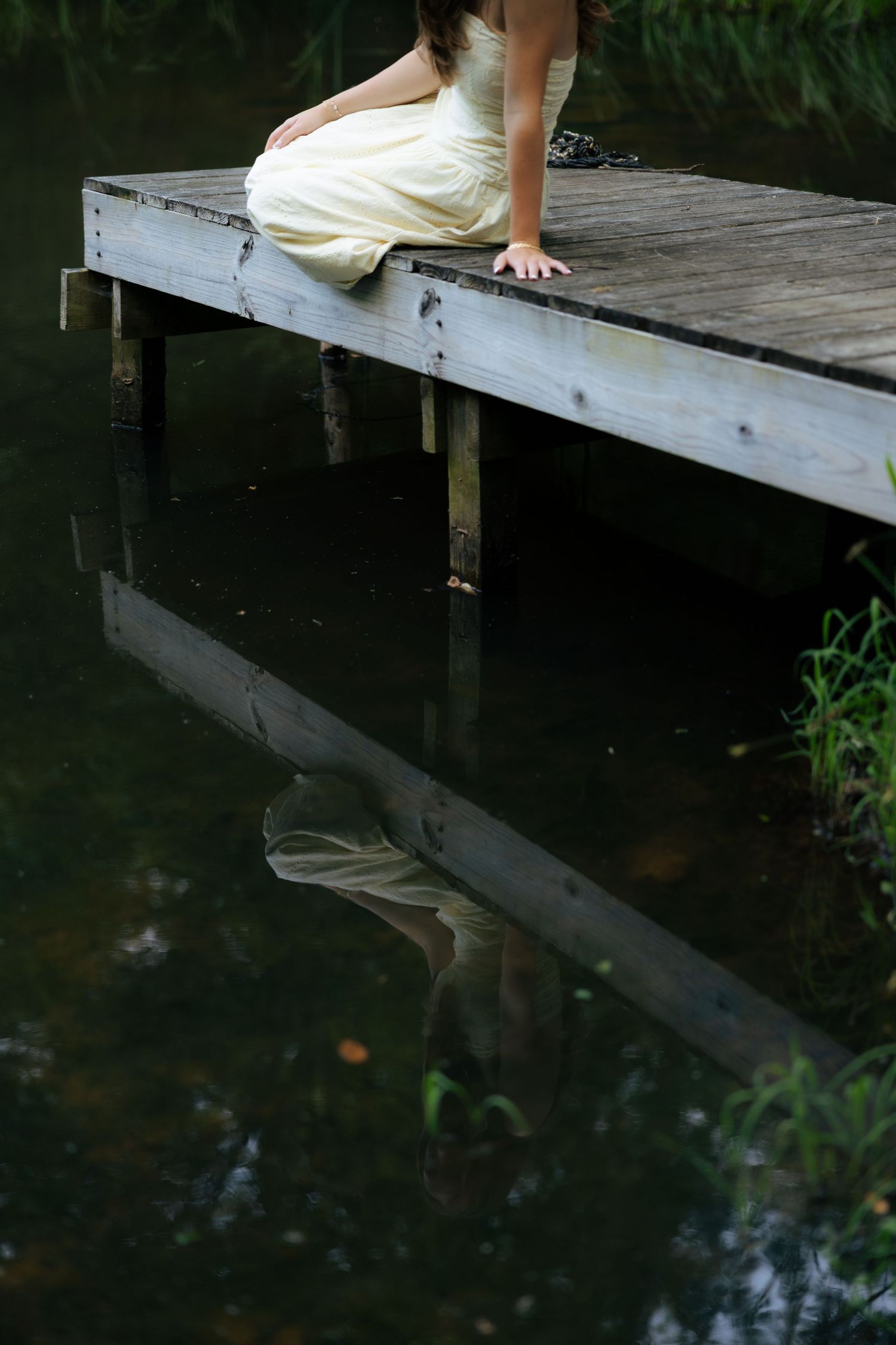 Young woman in yellow dress sitting on wooden dock reflected in dark still water.