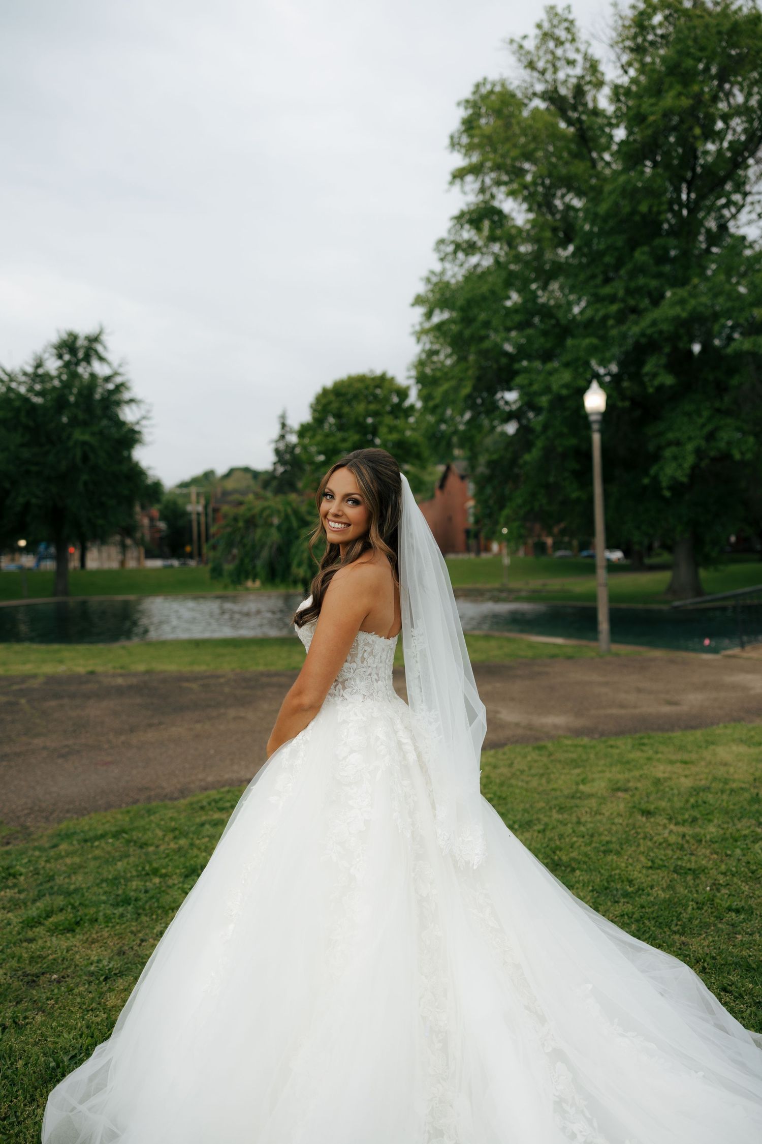 Smiling bride in a strapless white ballgown with veil poses outdoors in a lush green park setting.