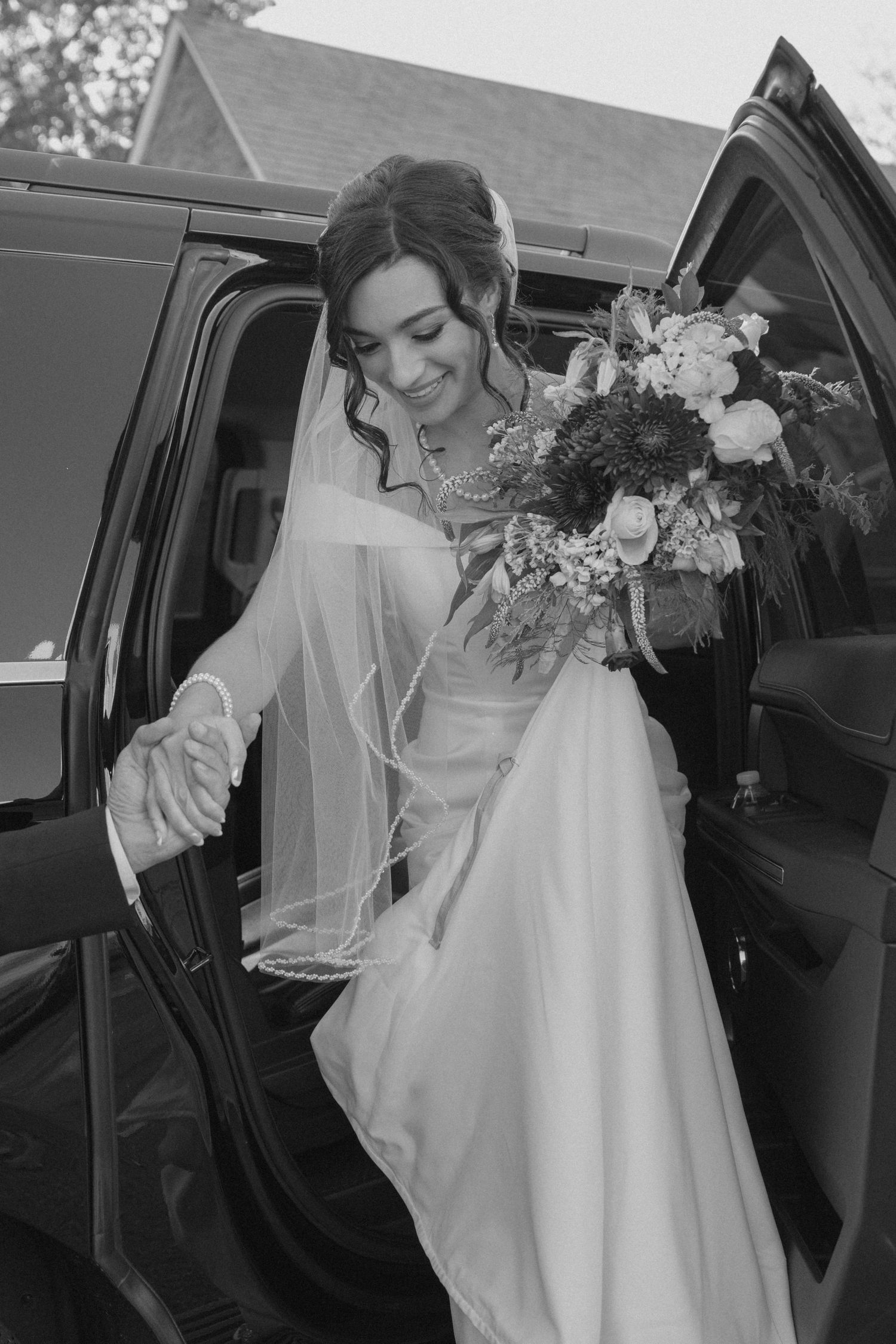 Bride in white gown and veil holding bouquet stepping out of black car on wedding day.
