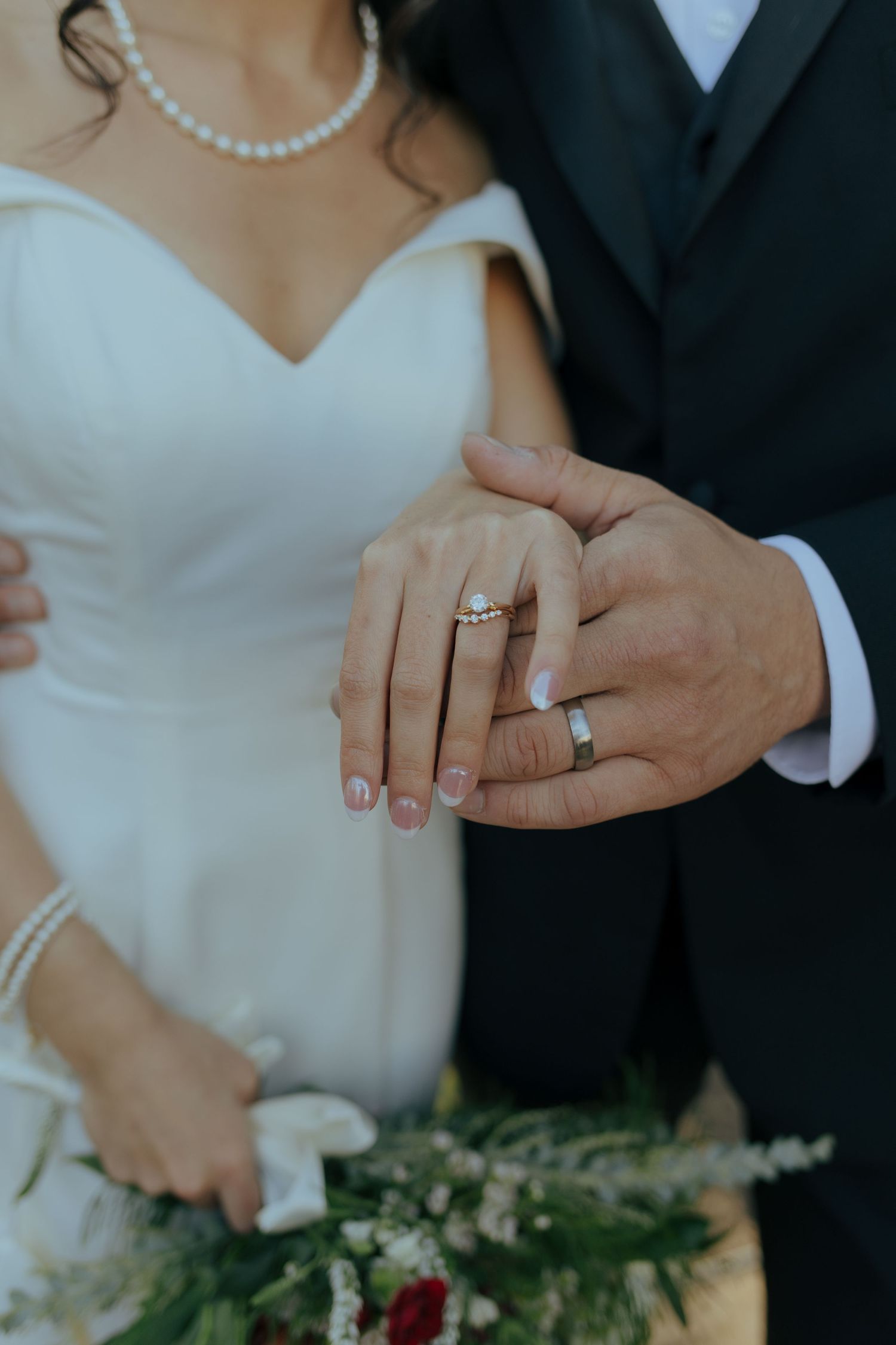 Bride and groom display their wedding rings, with the bride holding a floral bouquet.