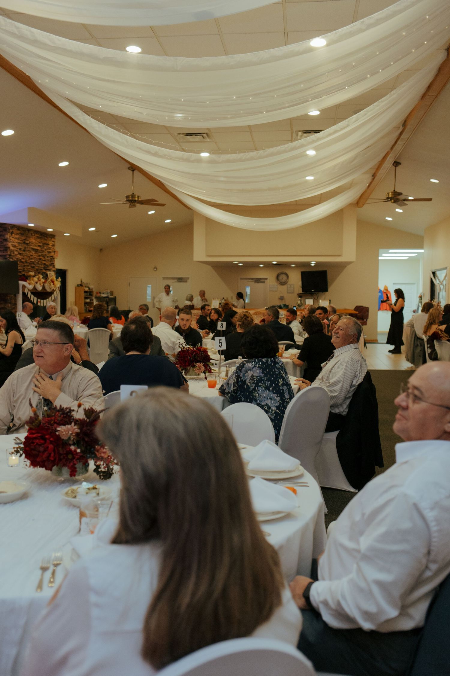 Wedding reception hall filled with guests seated at round tables under elegant white draped ceiling decor.