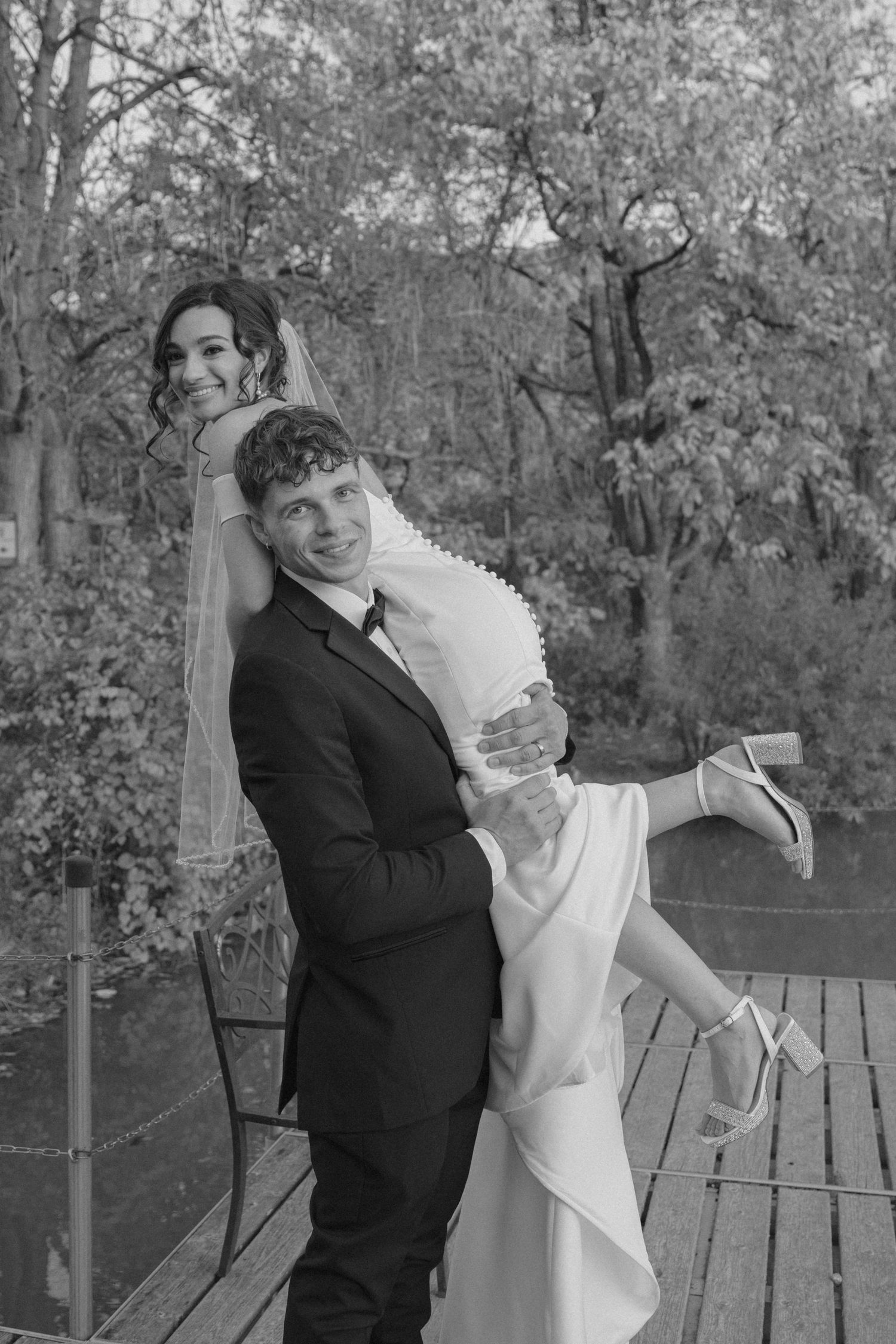 Groom joyfully carries smiling bride in his arms on a wooden bridge surrounded by lush autumn trees.