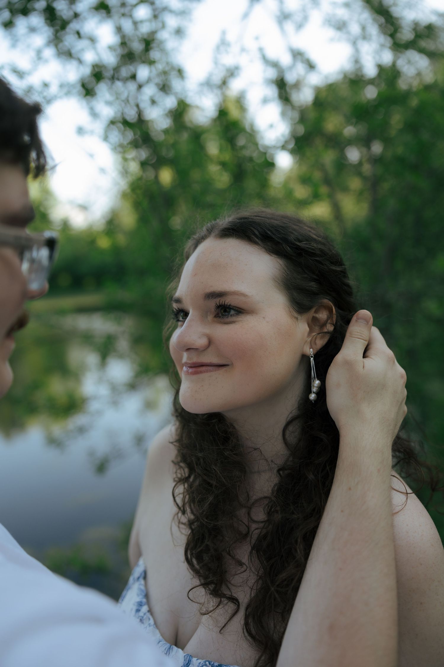 Young woman with curly hair smiling as her partner gently tucks hair behind her ear near a pond.
