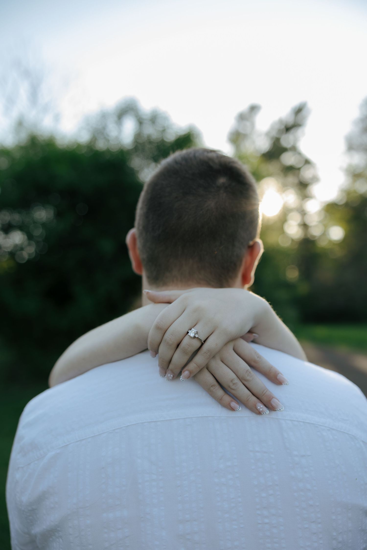 Woman's hand with diamond engagement ring draped over man's shoulder during outdoor engagement photo session.