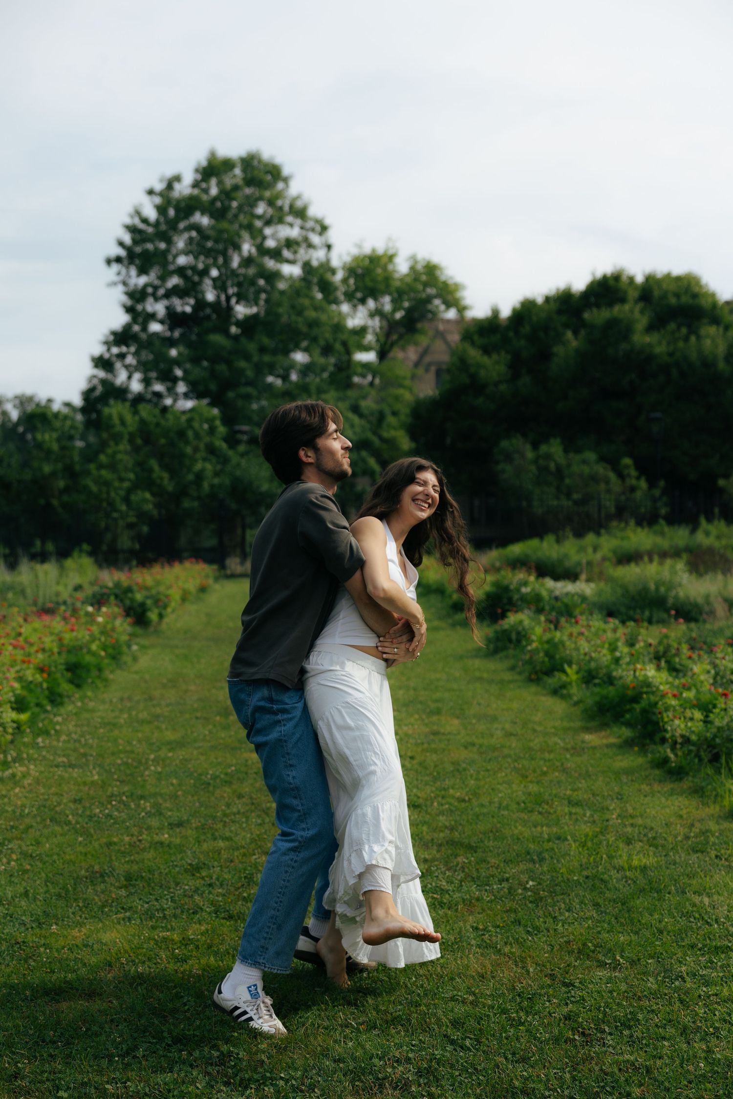 A couple embraces on a green lawn, the woman in a white dress laughing as her hair blows in the breeze.