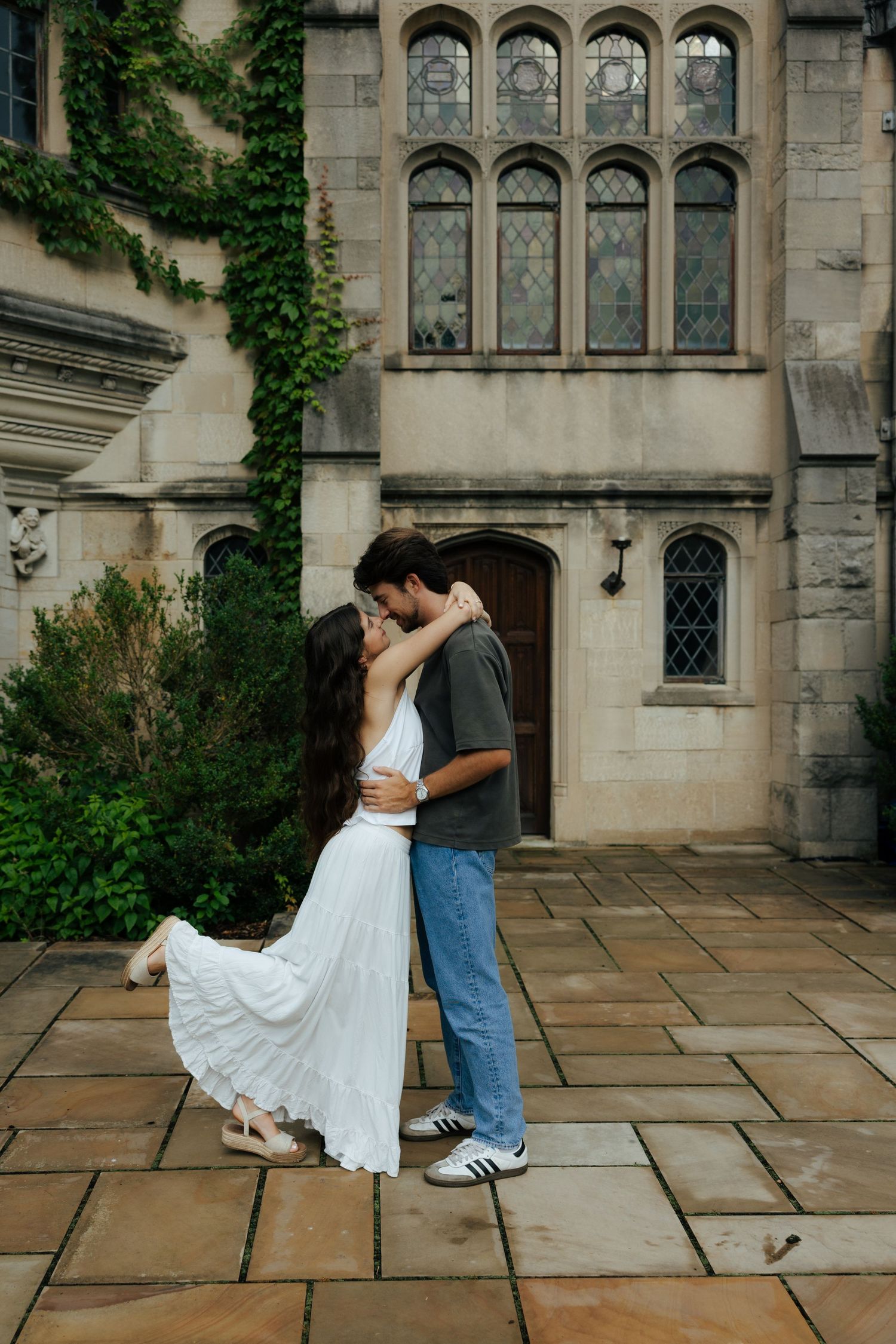 Couple sharing a romantic kiss in front of a gothic stone building covered in ivy on a cobblestone patio.