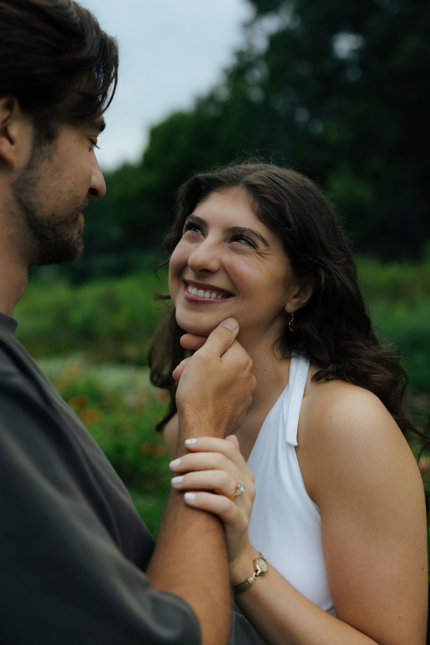 A smiling woman gazes up at a man who gently cups her chin in a lush green park setting.