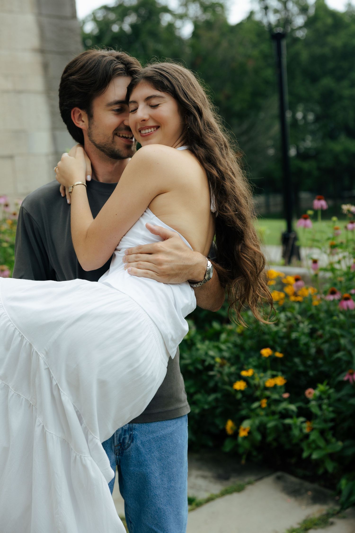 A smiling man carries a laughing woman in a white dress in his arms in a lush garden setting.