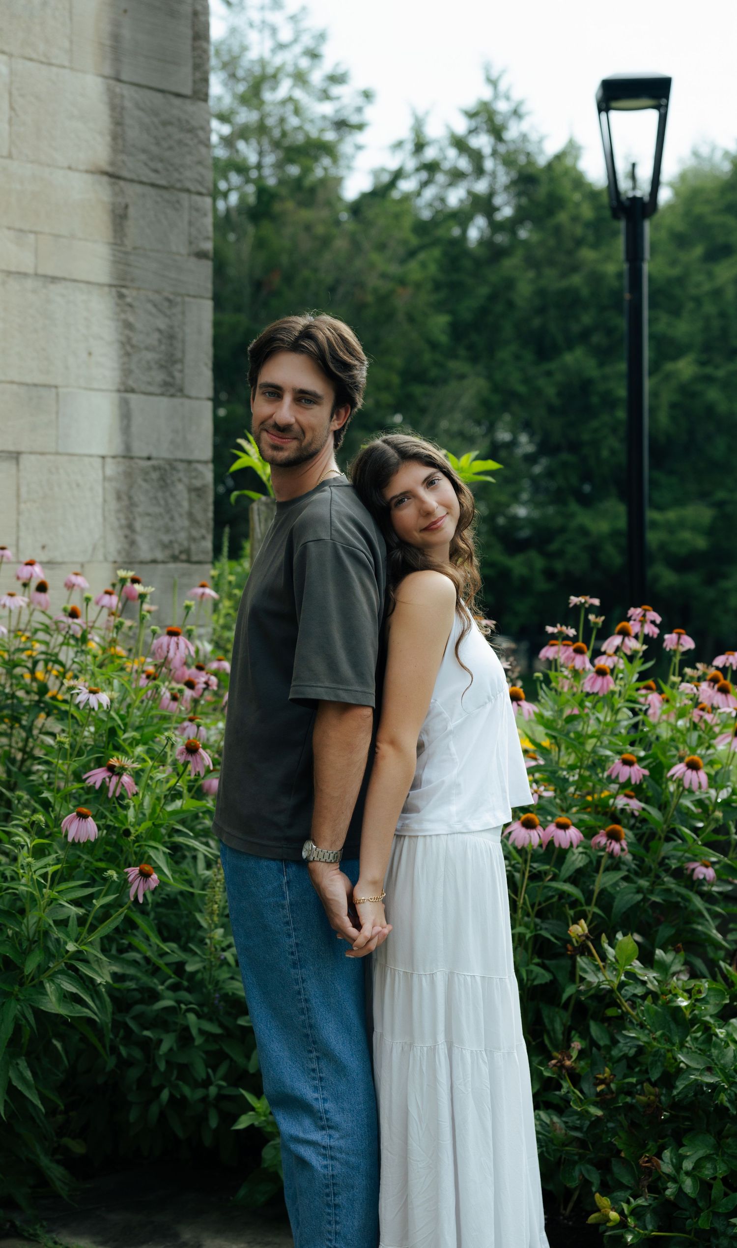 A couple holds hands and poses together in front of colorful pink flowers beside a stone pillar outdoors.