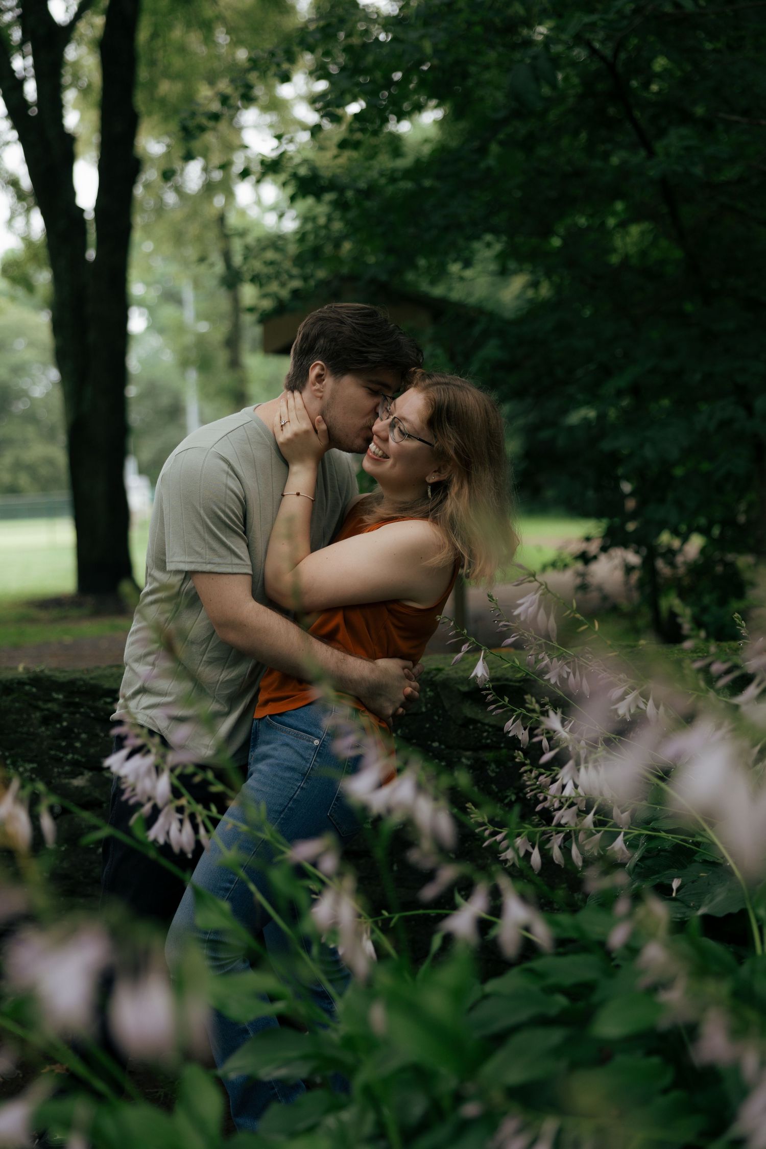 A couple shares a close romantic moment surrounded by blooming flowers and lush green trees in a park.