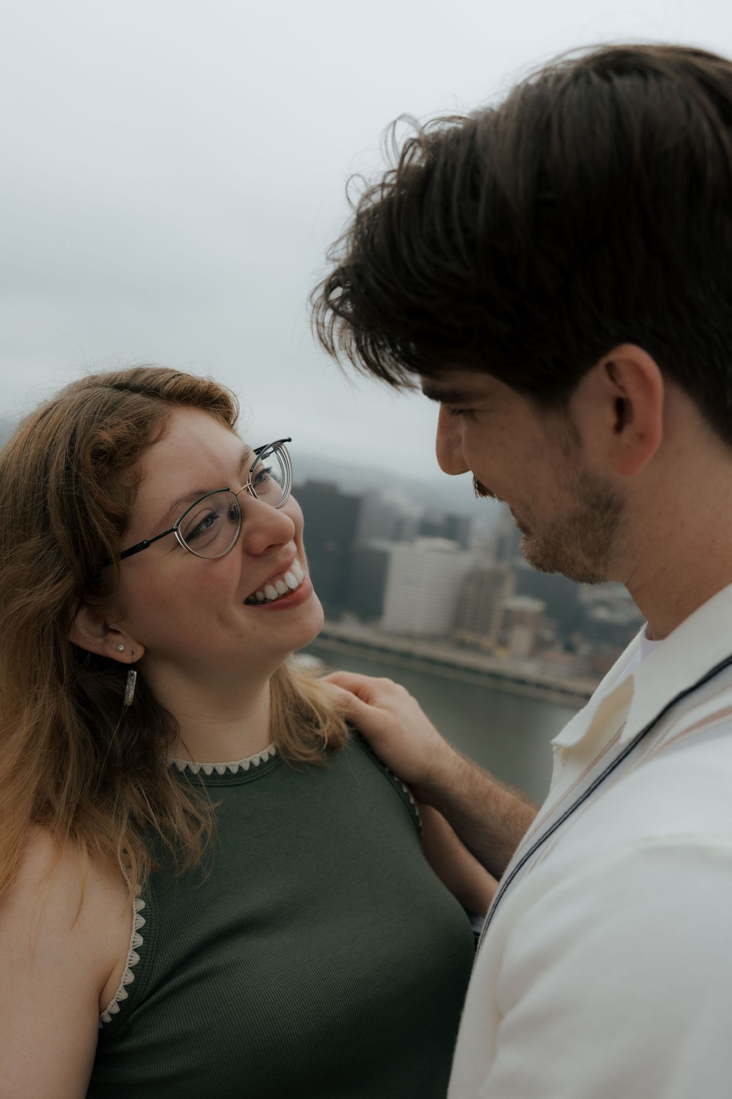 Smiling woman with glasses gazes at her partner during a romantic outdoor engagement session by the river.