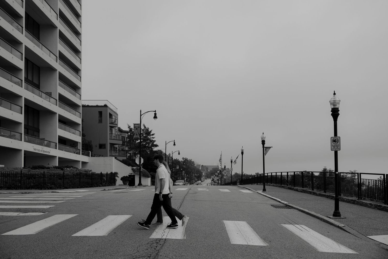 Black and white photo of two people crossing a zebra crossing on an empty urban street on a cloudy day.