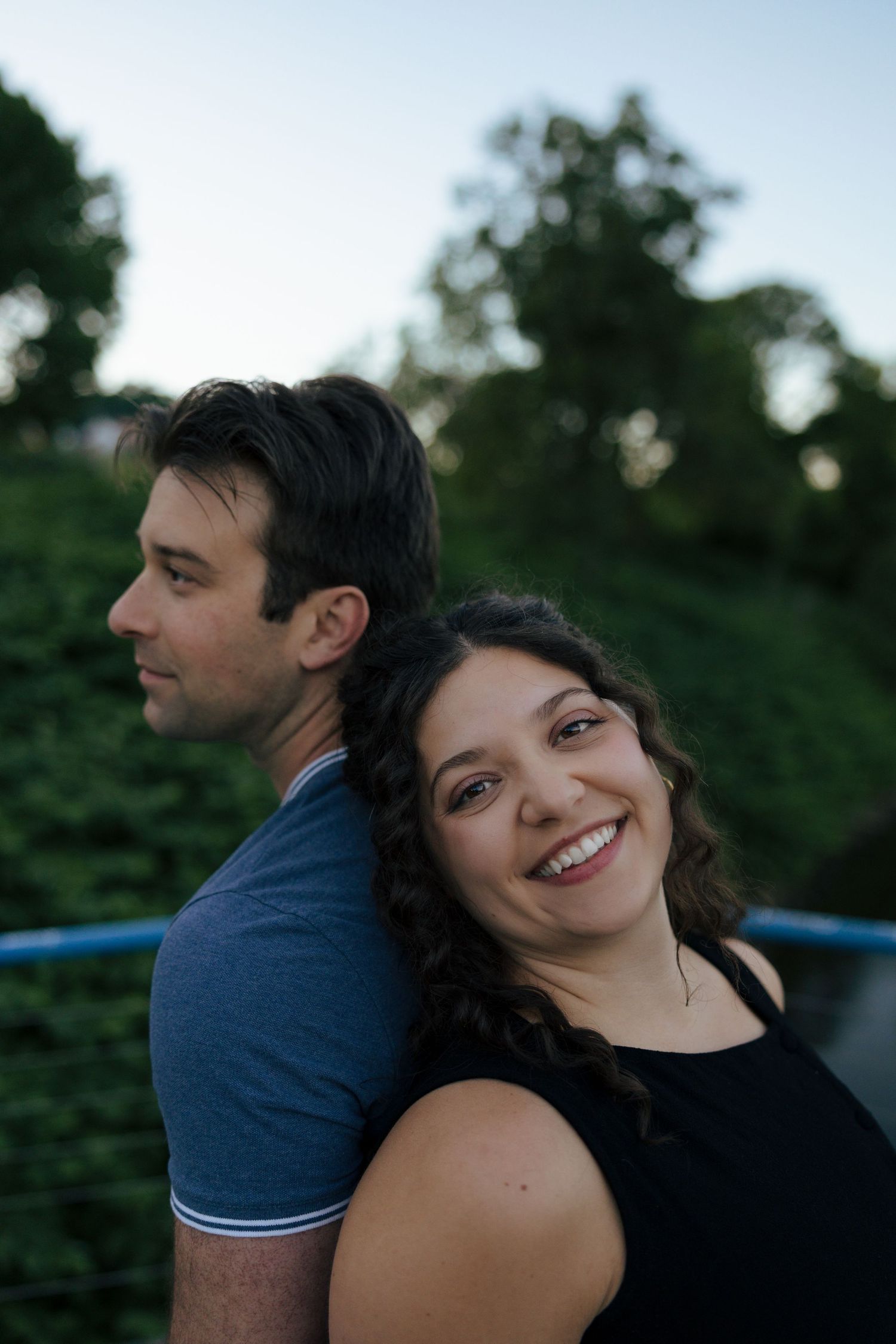Smiling young woman leans back-to-back with a man outdoors at dusk with green foliage behind them.