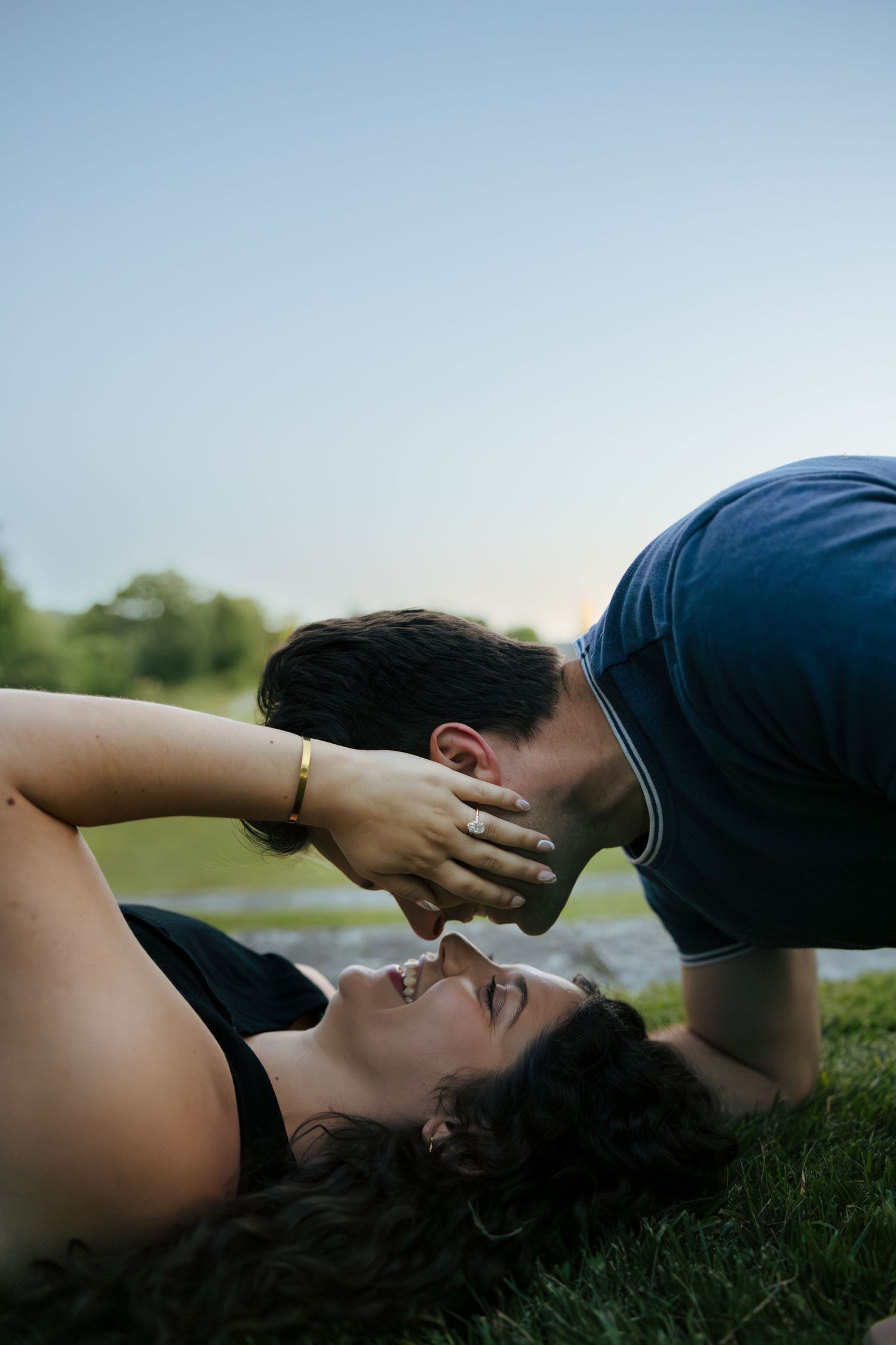 Man leaning down to kiss smiling woman lying on grass by a peaceful lakeside at sunset.
