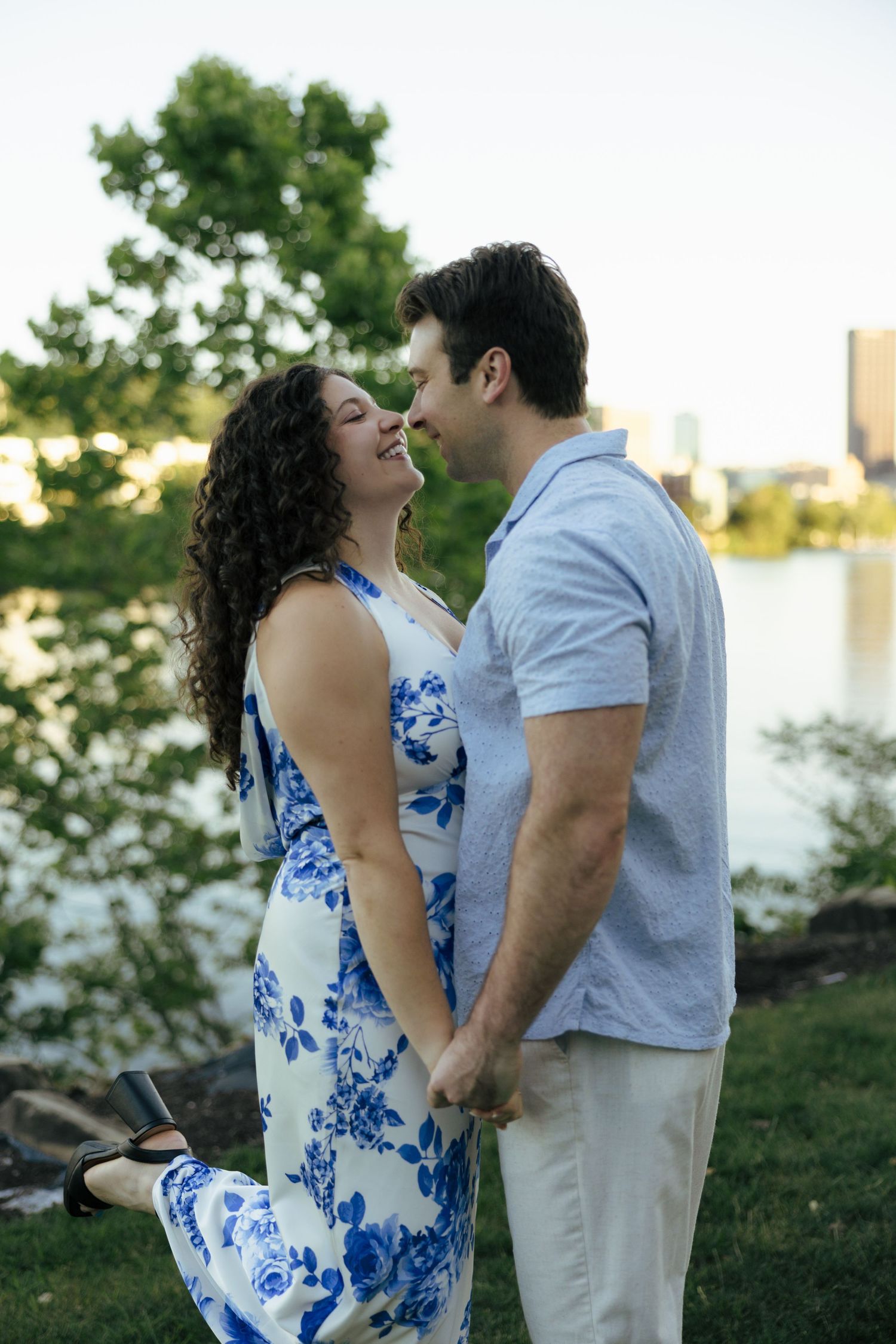 A couple holds hands and leans in for a kiss near a sunlit river with a city skyline.