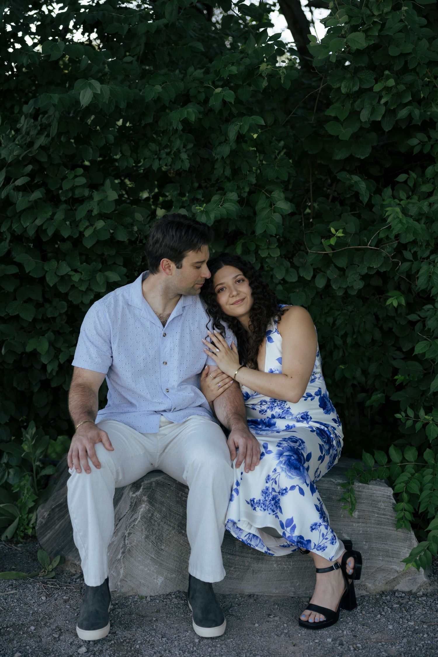 Engaged couple sitting on rock surrounded by lush greenery, sharing a tender romantic moment.