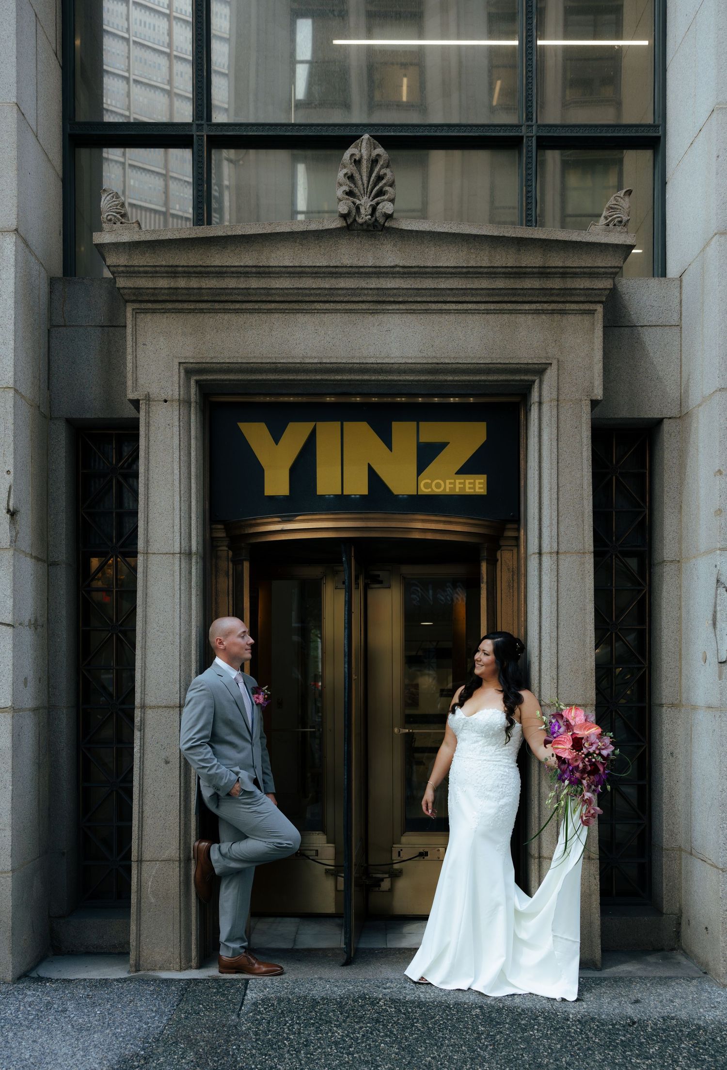 Bride and groom posing outside Yinz Coffee shop entrance, bride in white gown holding bouquet, groom in gray suit.