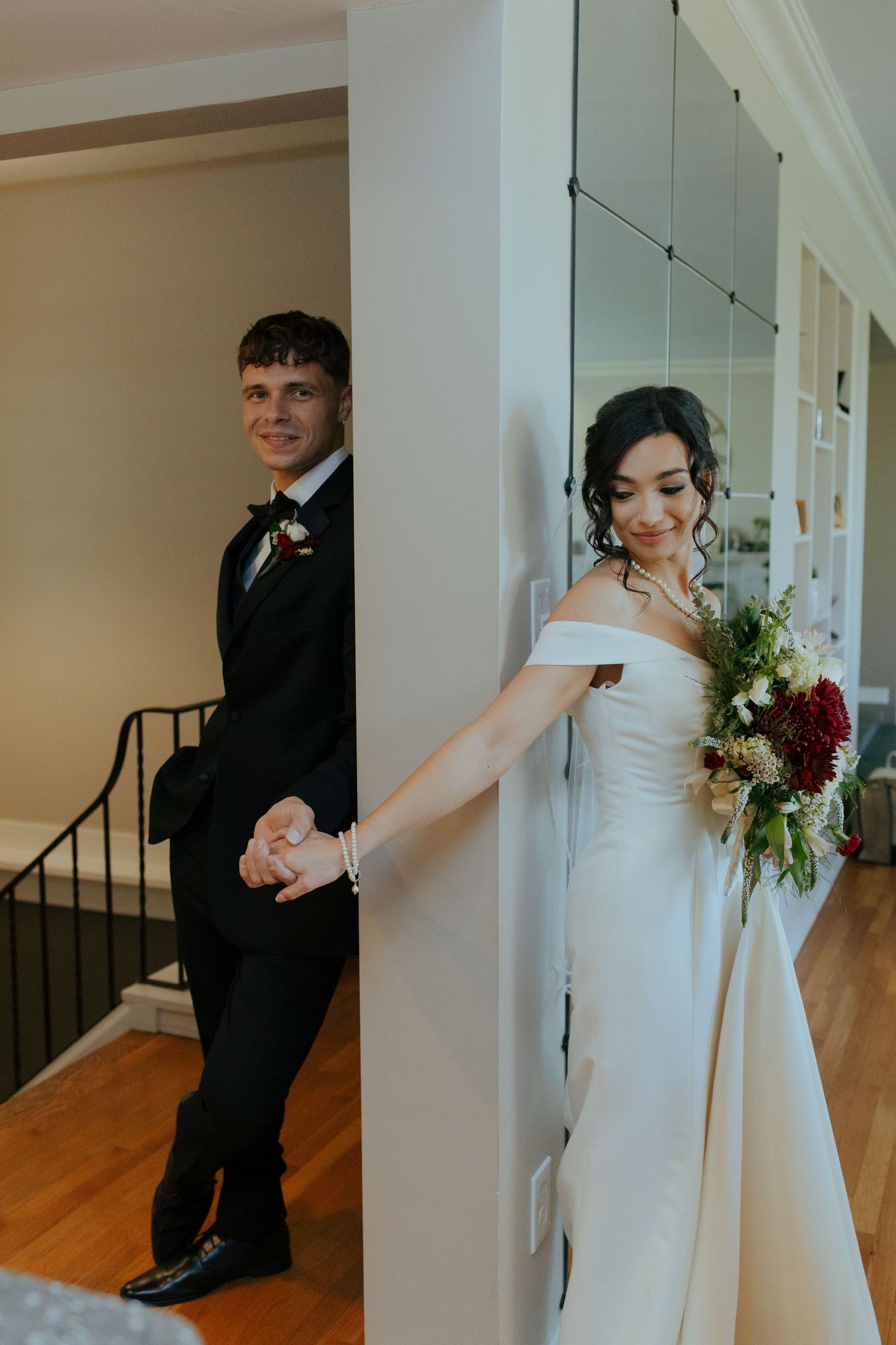 Bride and groom hold hands around a white column during first look, bride holding a lush floral bouquet.