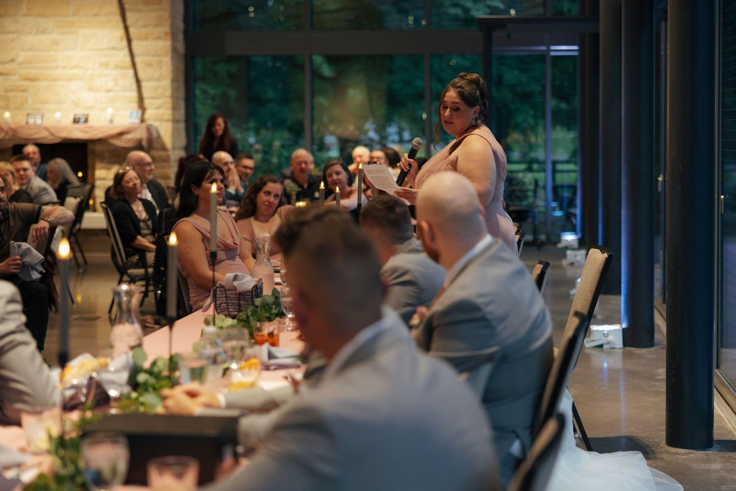 A woman gives a toast speech at an elegant indoor wedding reception dinner with guests seated at decorated tables.