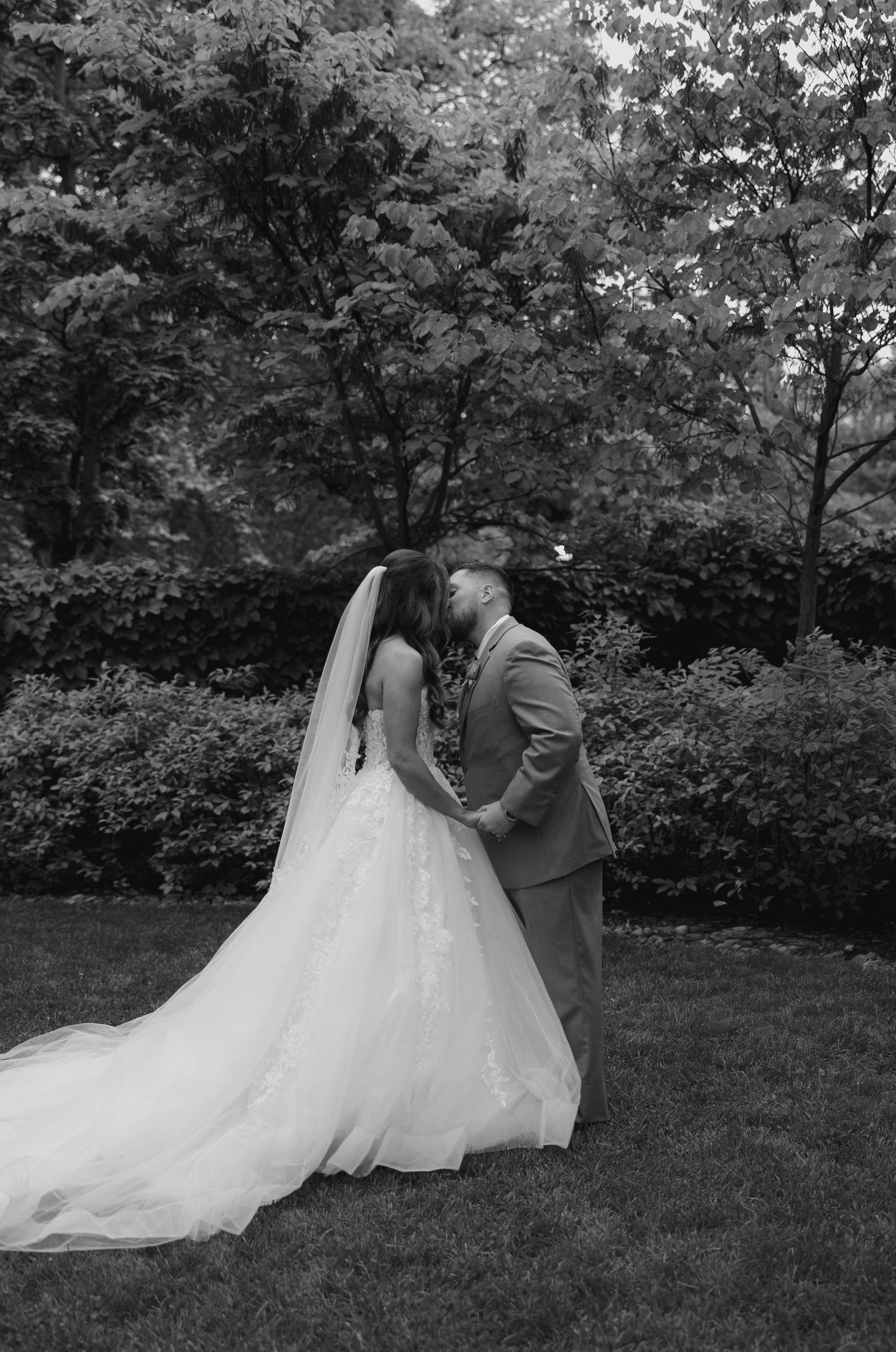 Black and white photo of bride and groom sharing a kiss in lush garden after wedding ceremony.