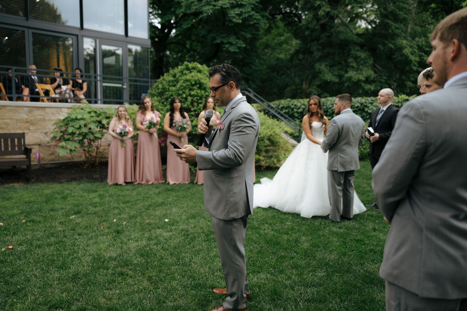 Groomsman reads speech during outdoor wedding ceremony with bridesmaids in pink dresses and bride in white gown on green lawn.