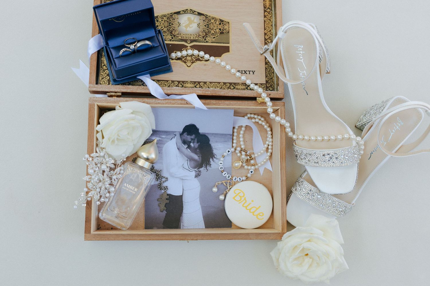 Wedding flat lay with bridal heels, pearl jewelry, rings, photo, white roses, and perfume in a wooden box.