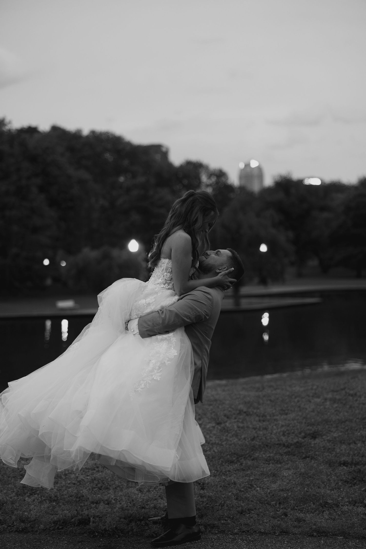 Groom lifts bride in flowing white ball gown near a serene lake at twilight in black and white.