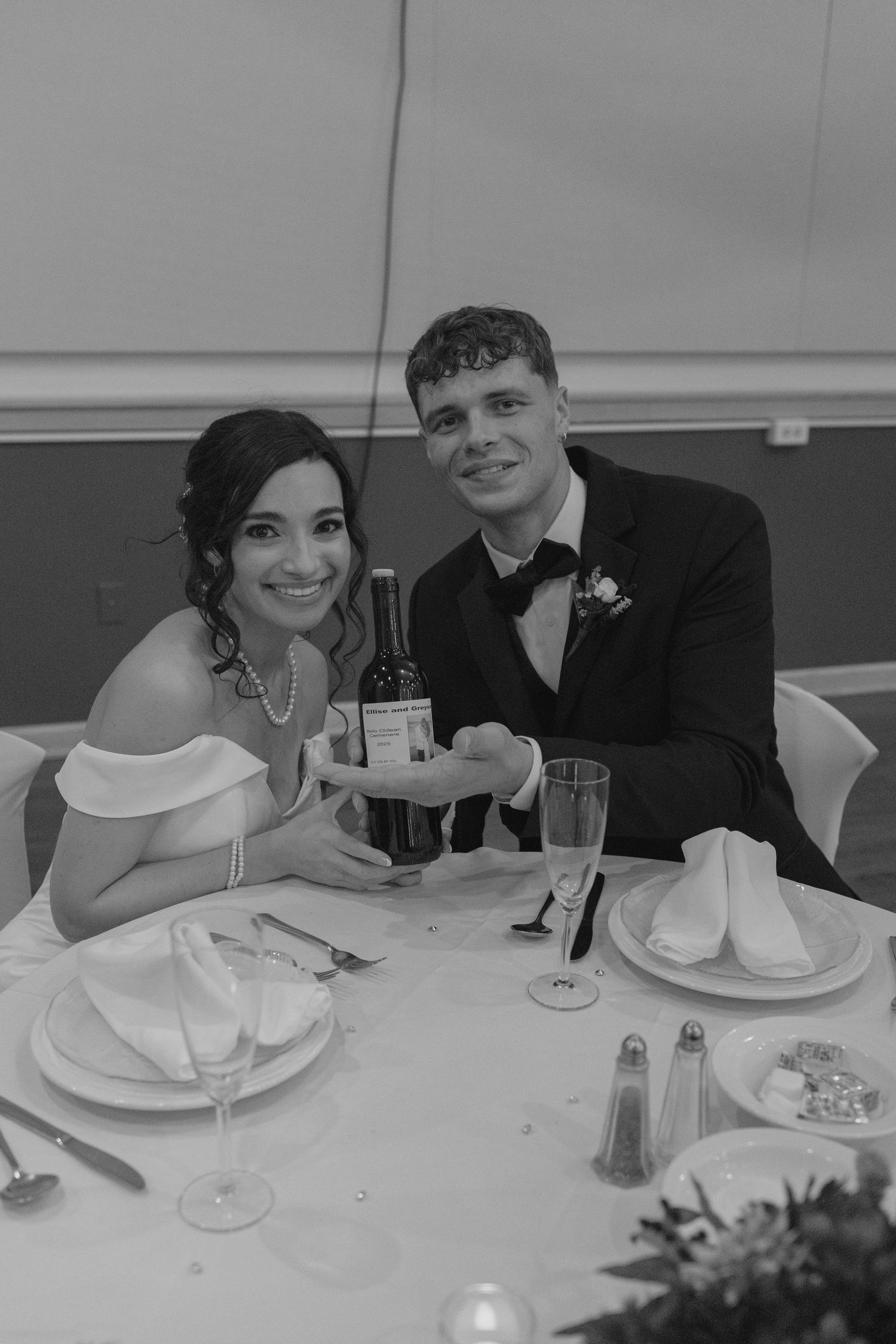 Bride and groom smiling at wedding reception table holding wine bottle in black and white photo.