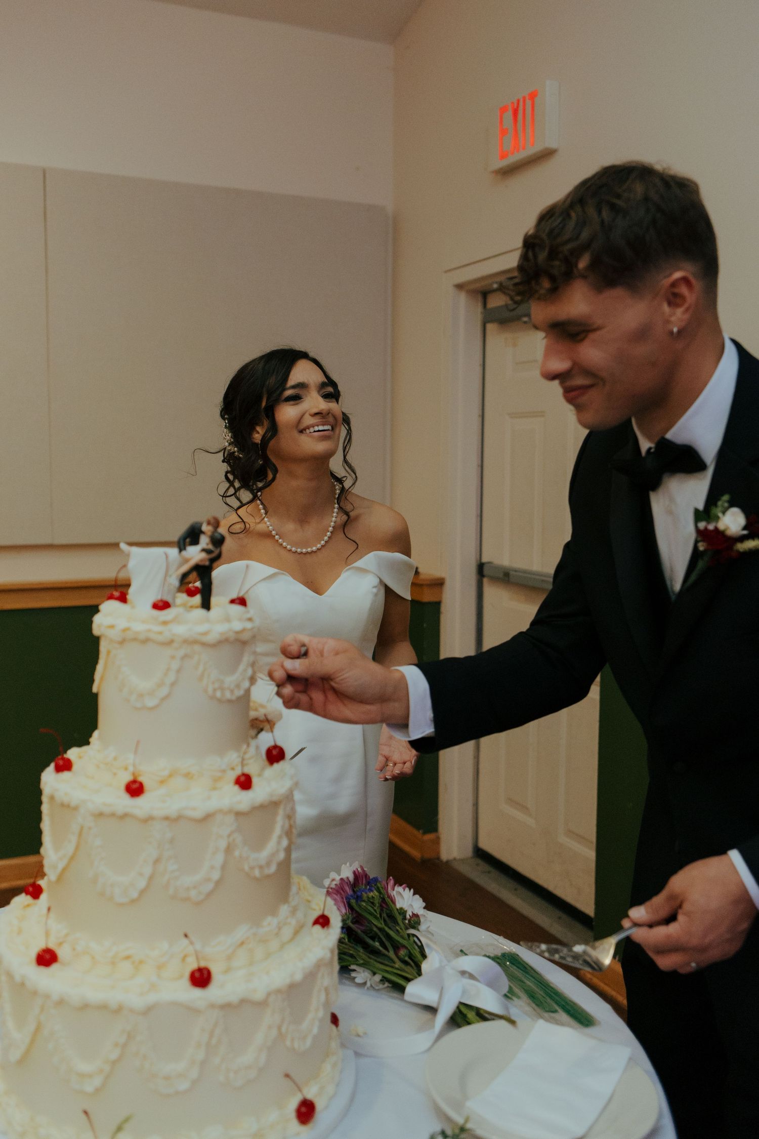 Smiling bride and groom cutting a three-tier white wedding cake decorated with cherries at their reception.