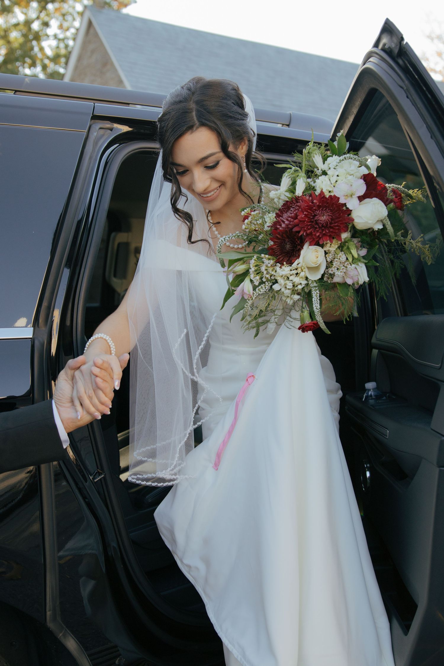 Elegant bride in white gown holding a red and white bouquet stepping out of a black luxury vehicle.