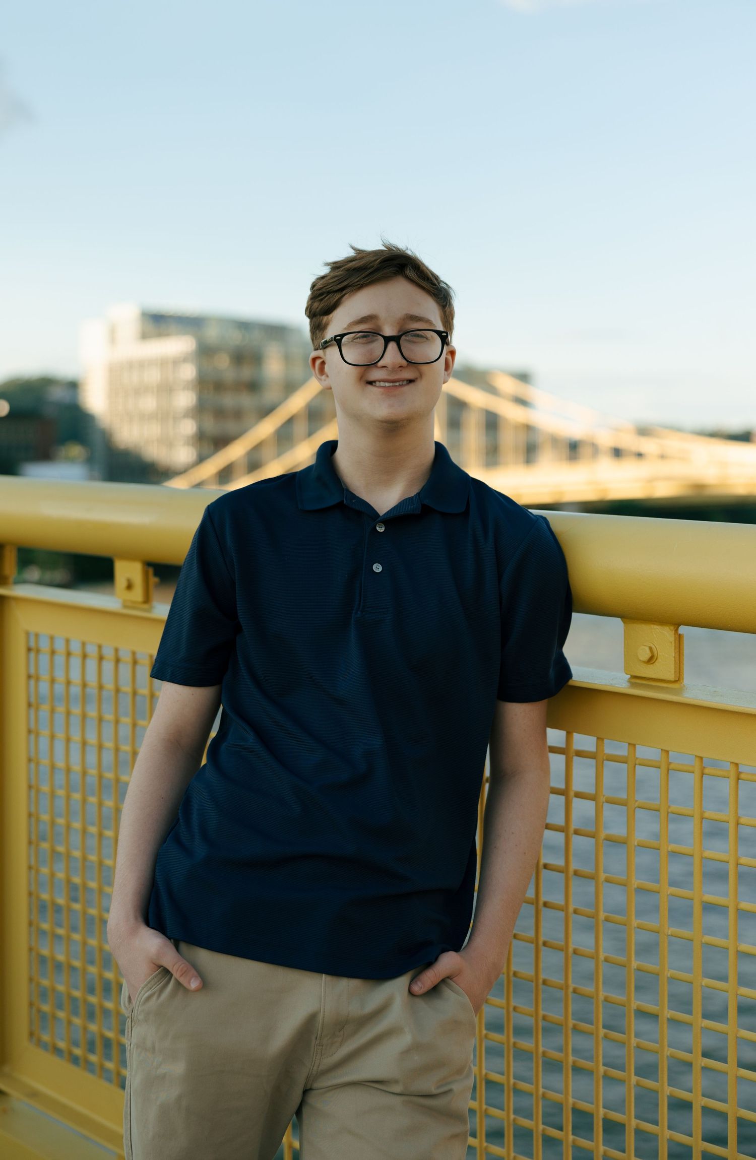 Smiling teenage boy in navy polo leaning on yellow bridge railing with Pittsburgh skyline behind him.