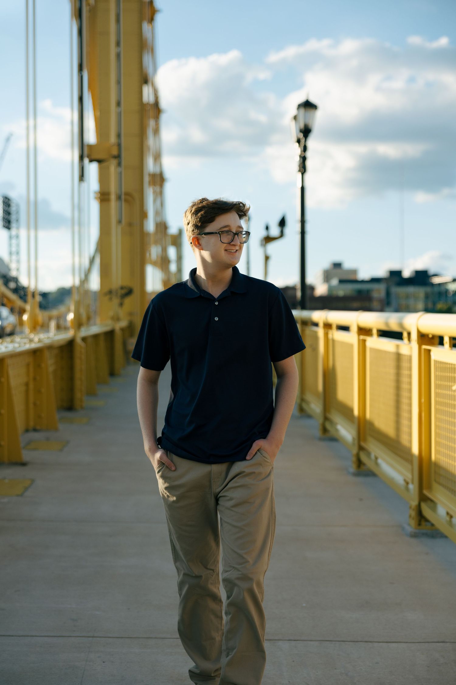 Teen boy in navy polo and khakis walks on Pittsburgh's yellow Roberto Clemente Bridge at golden hour.