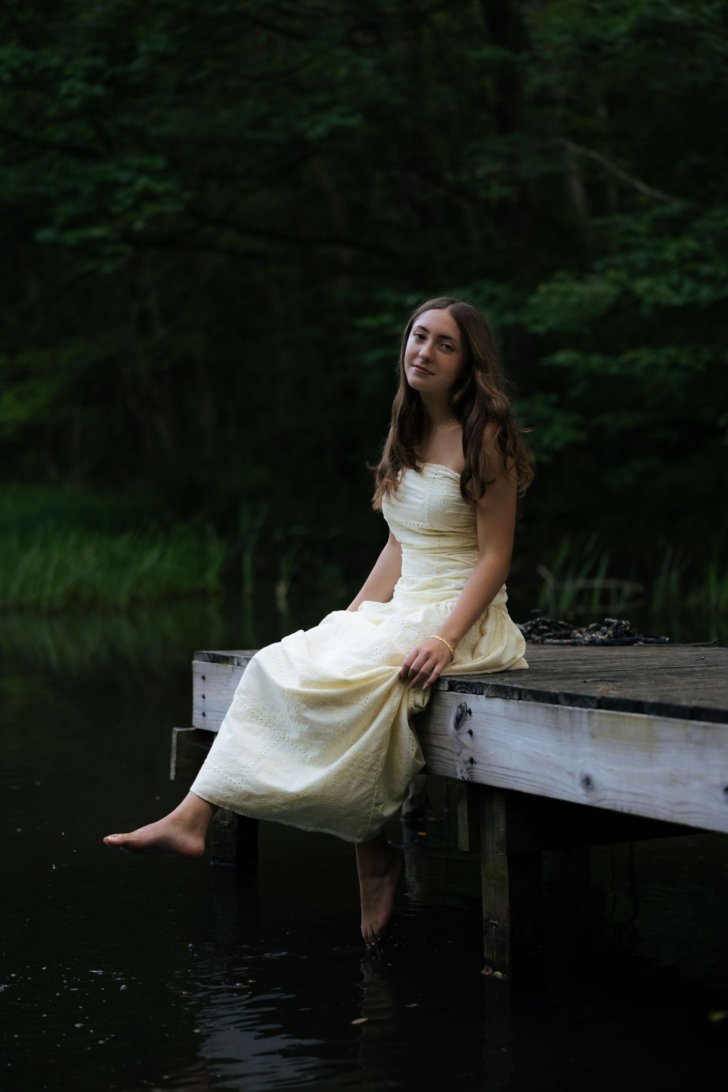Young woman in pale yellow strapless dress sitting on wooden dock edge over dark lake water.