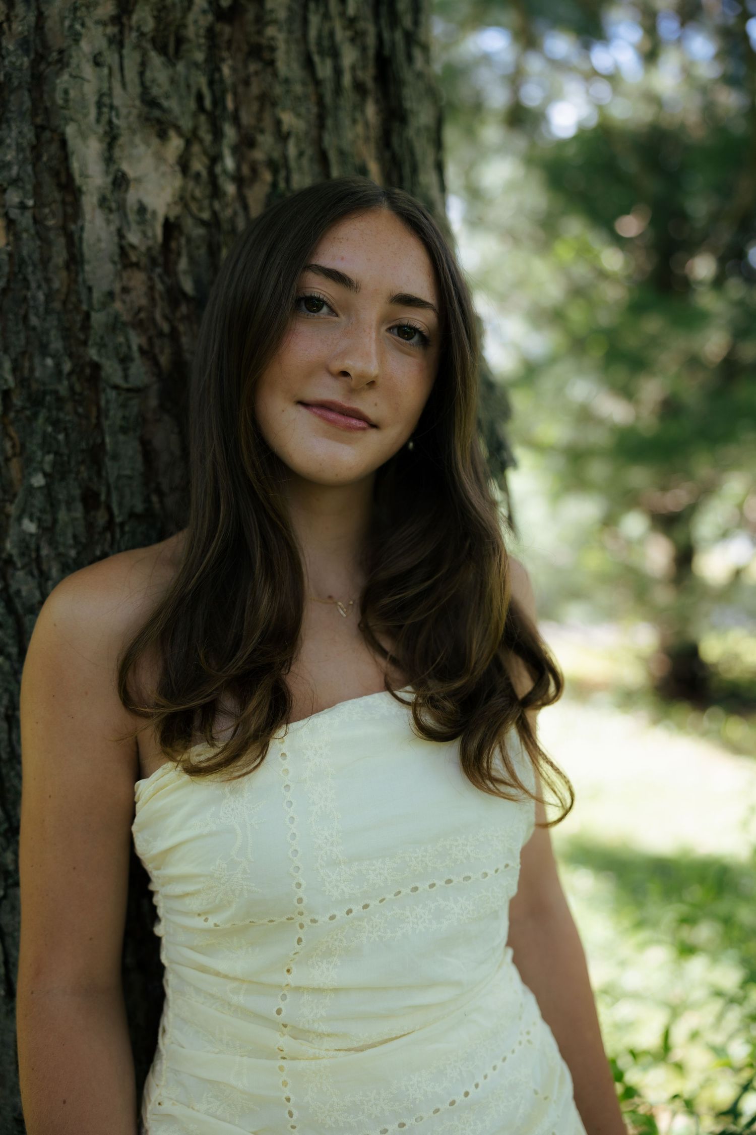 Young brunette woman in a strapless yellow dress leaning against a tree trunk in a sunny park.