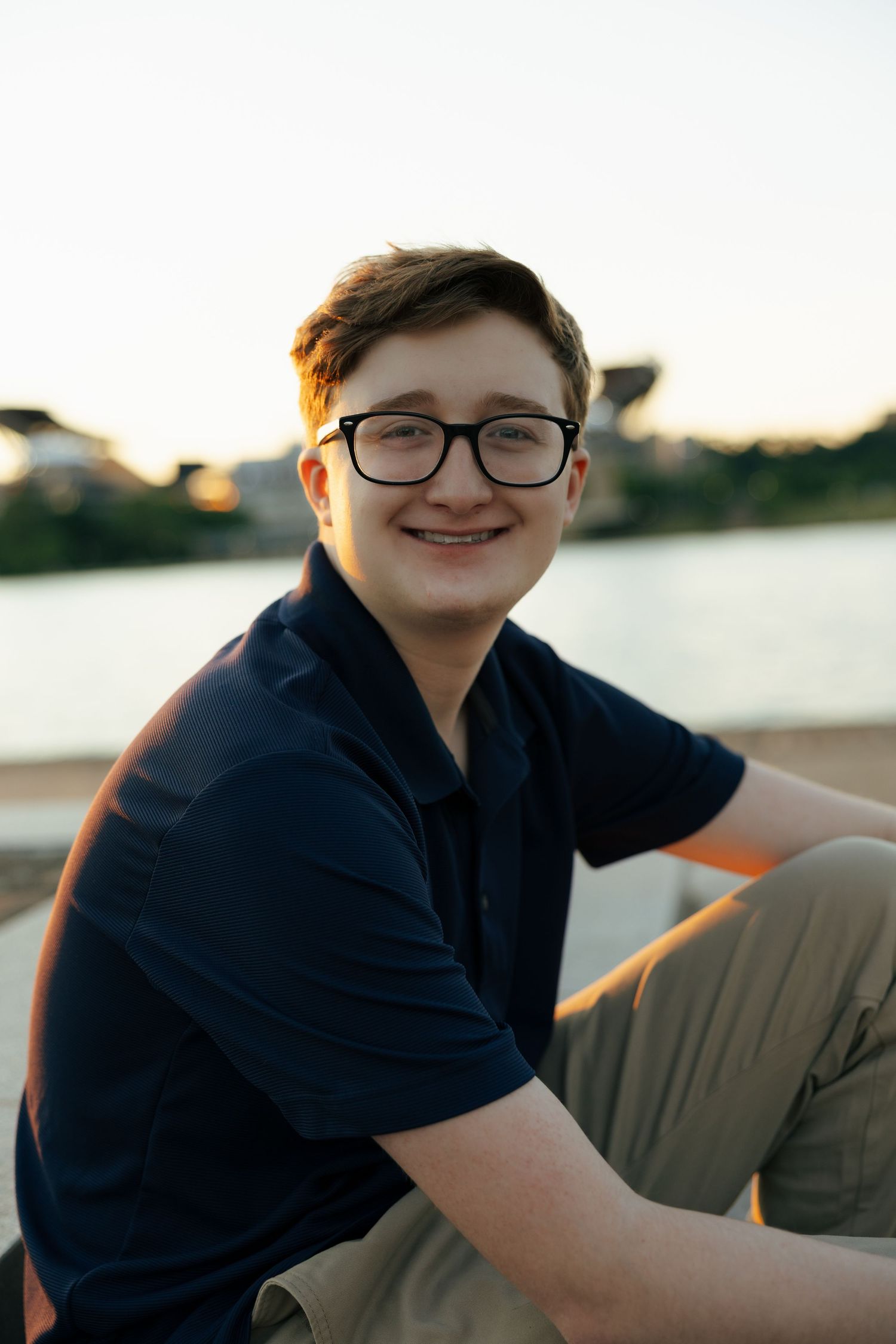 Teenage boy in navy polo and khakis sitting outdoors near waterfront, smiling at camera at sunset.