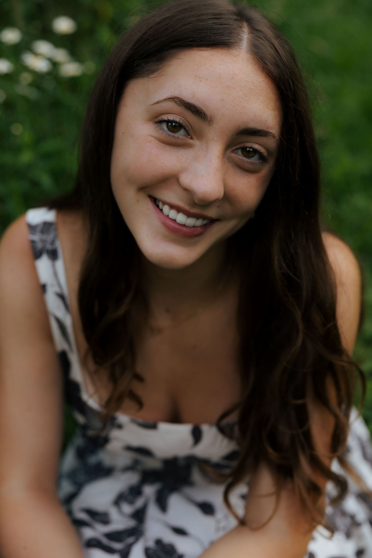 Close-up portrait of smiling brunette woman in floral dress with green foliage background.