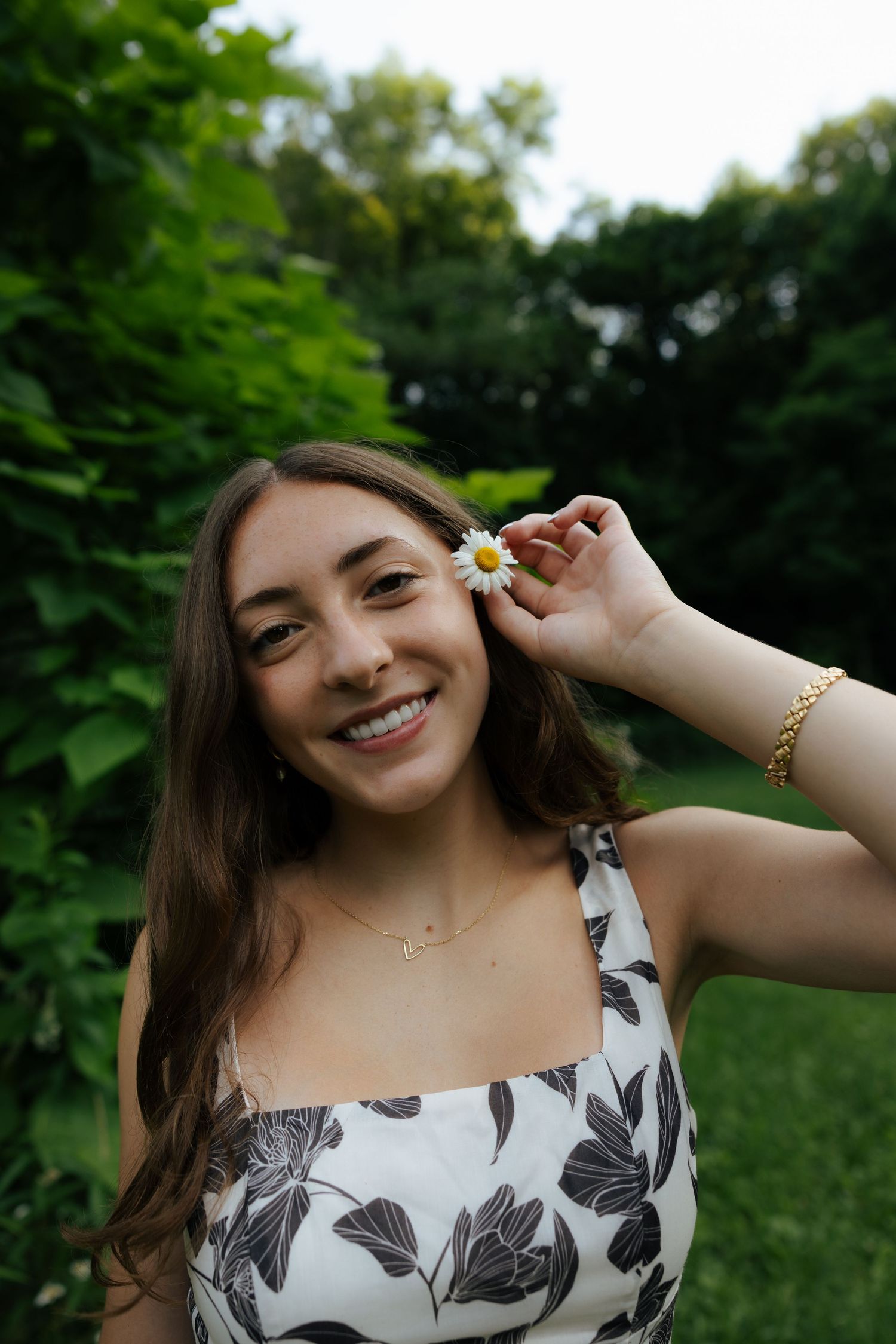 Smiling brunette woman holding daisy flower near ear, wearing floral dress and gold bracelet outdoors.