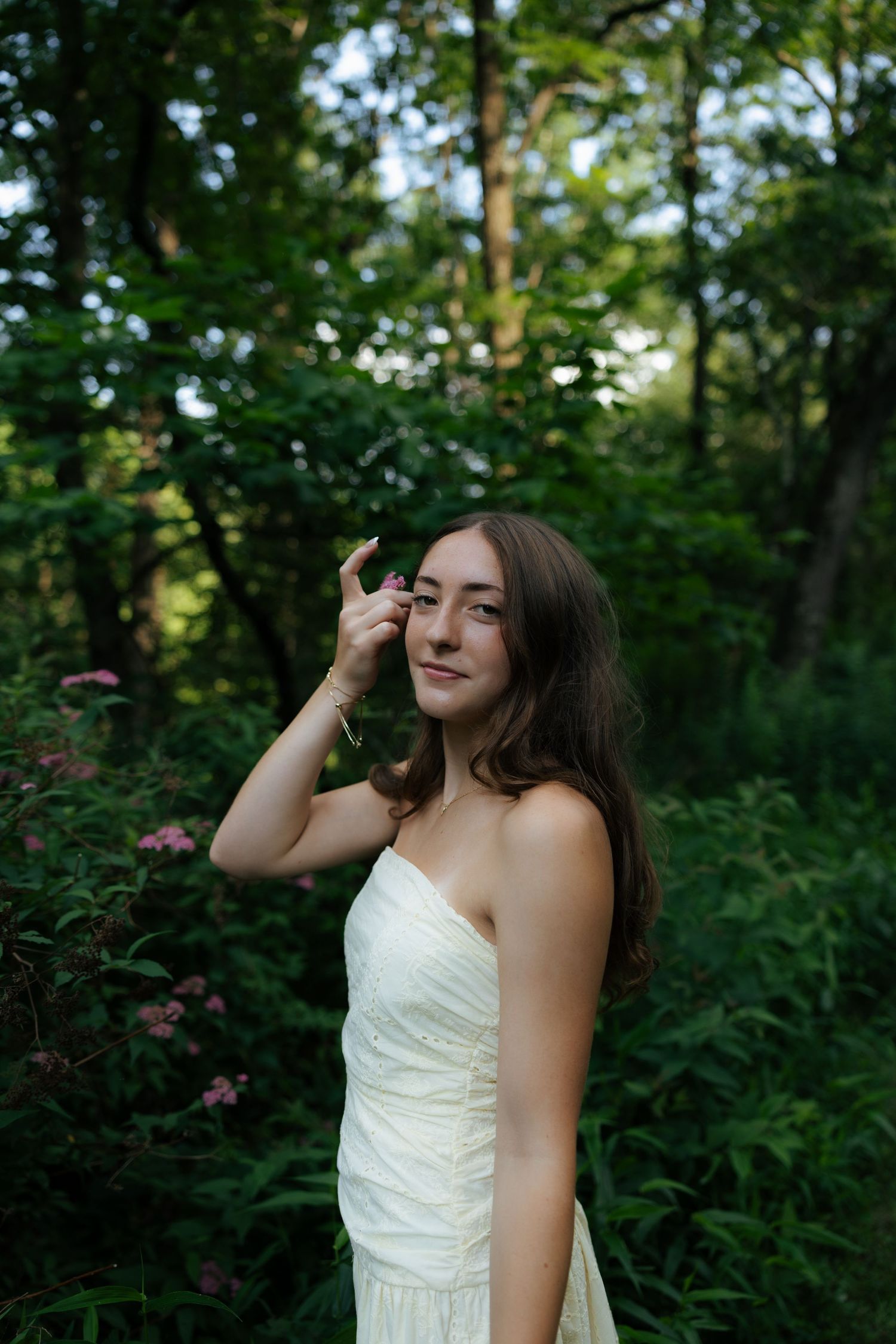 Young brunette woman in a white strapless dress standing among lush green trees and pink flowers in a forest.