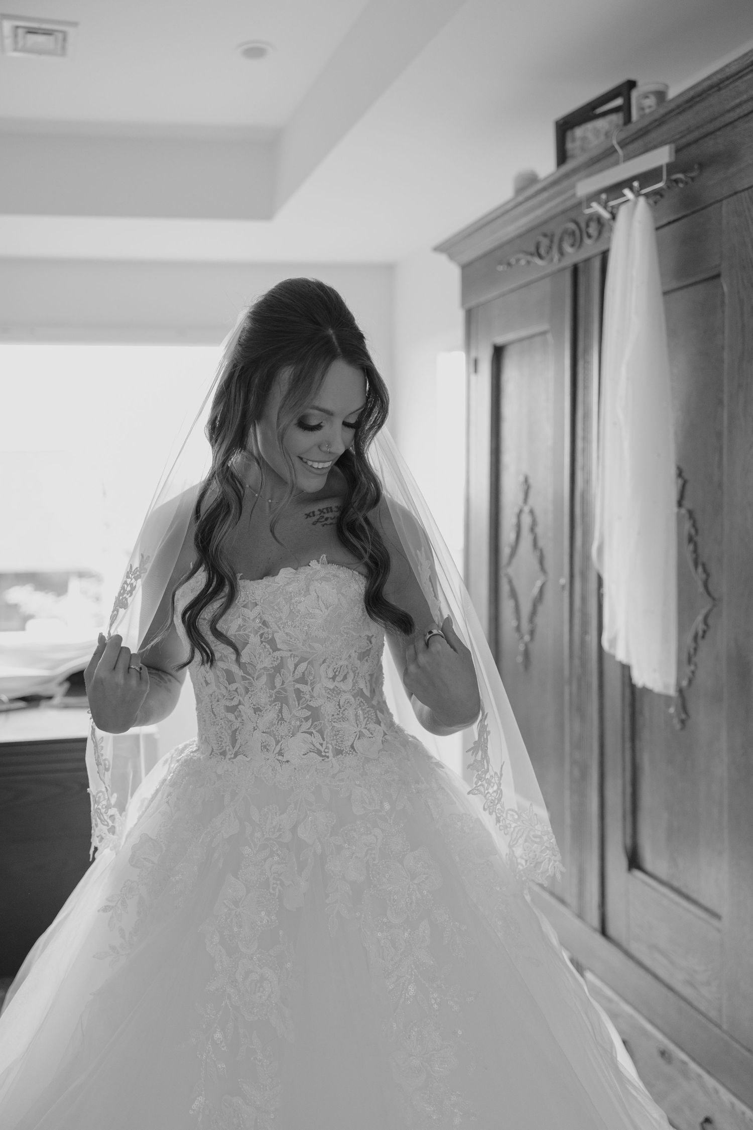 Black and white photo of a smiling bride in a lace ballgown and veil, standing near a wooden armoire.