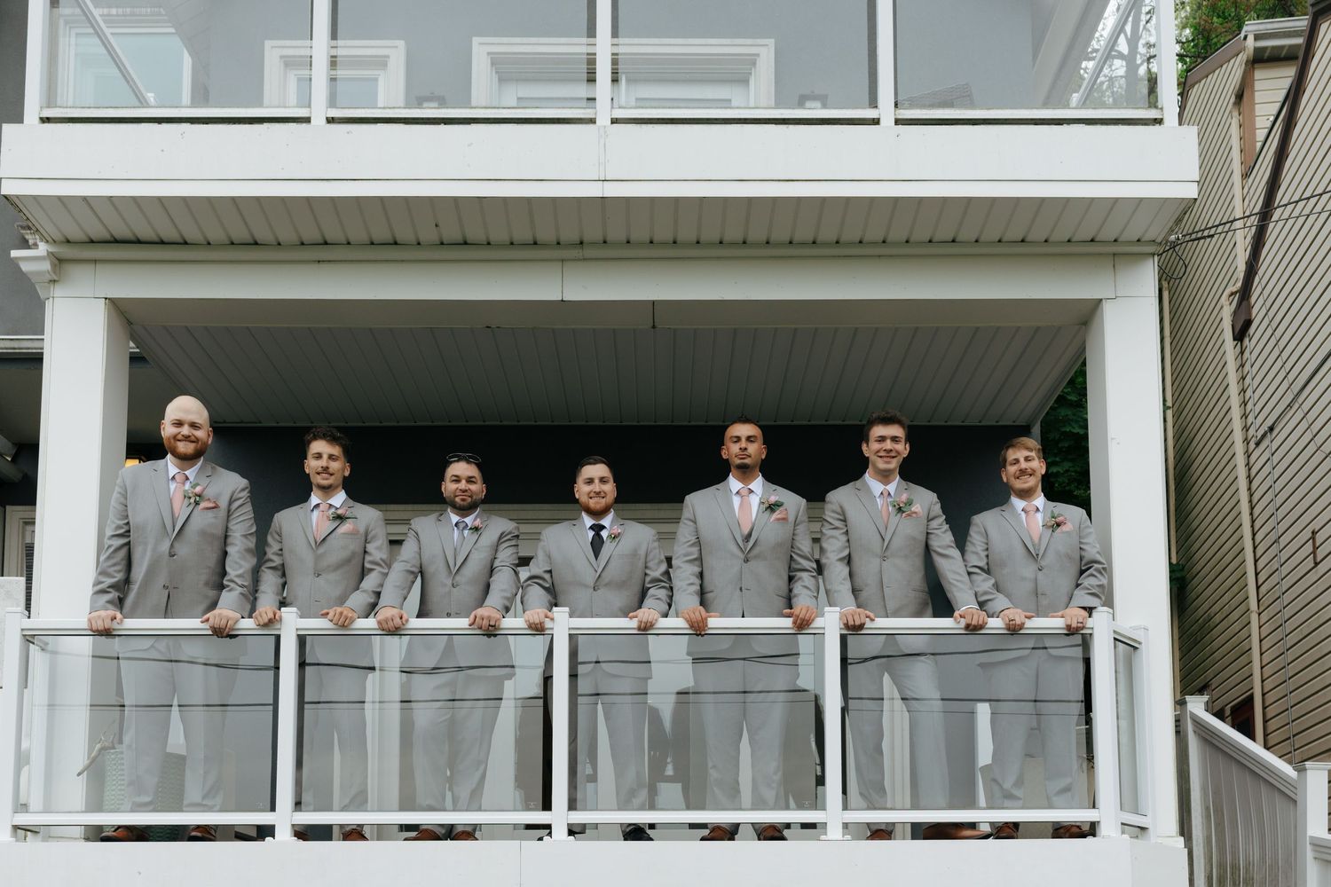 Seven groomsmen in gray suits with pink ties pose on a white balcony for a classic wedding party photo.