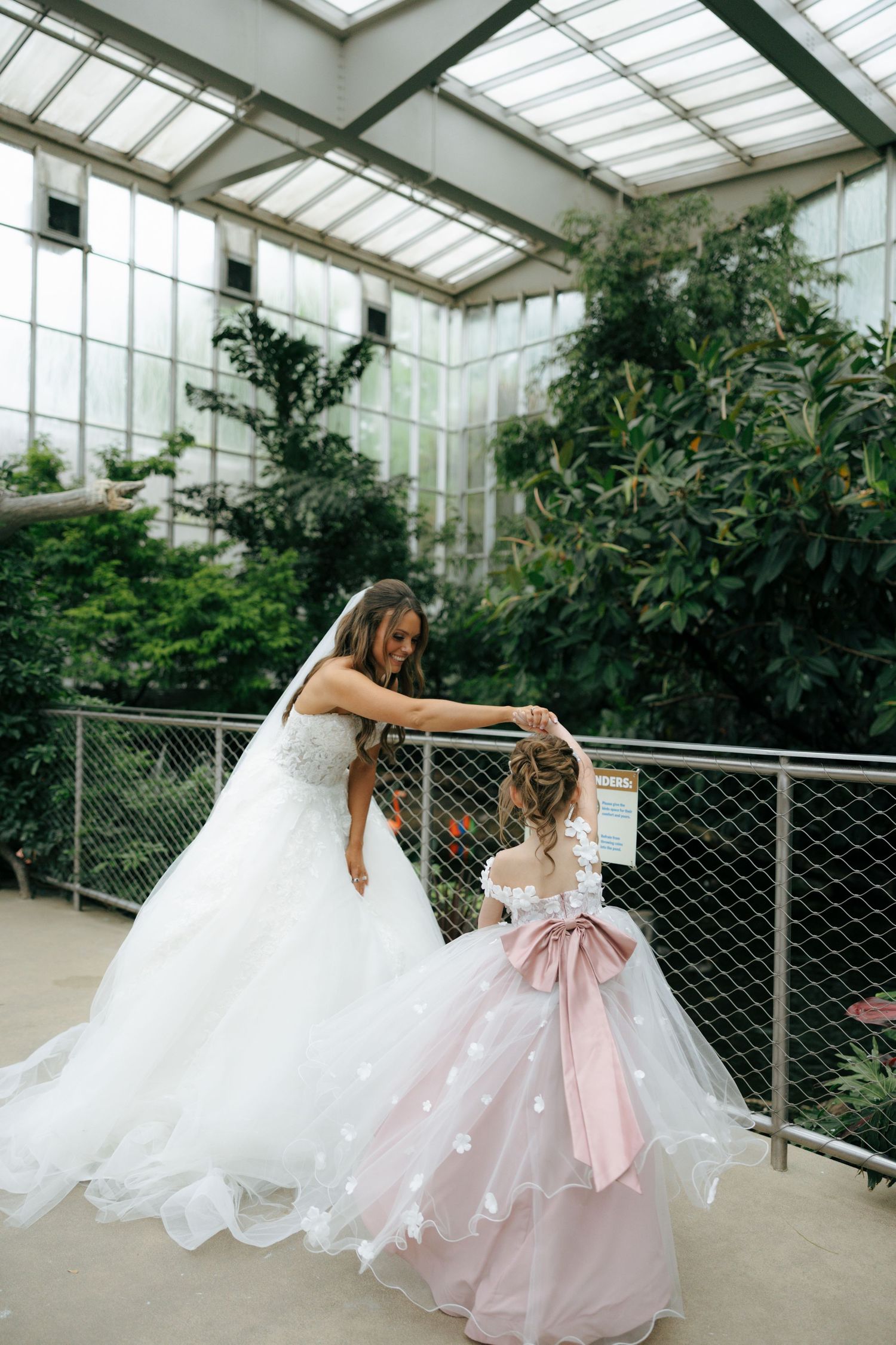 Bride in white gown adjusting flower girl's pink dress inside a lush greenhouse venue.